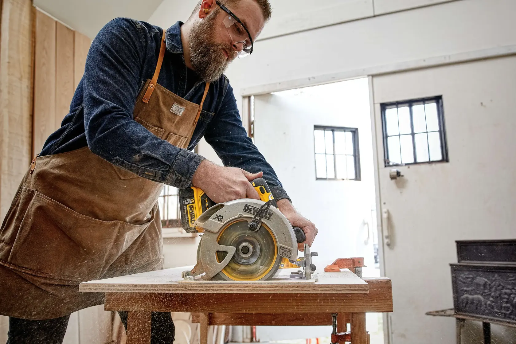 A person wearing a work apron and dark shirt is using a DEWALT cordless circular saw to cut a piece of wood in a workshop. The saw is yellow and silver, and sawdust is visible from the cutting process.
