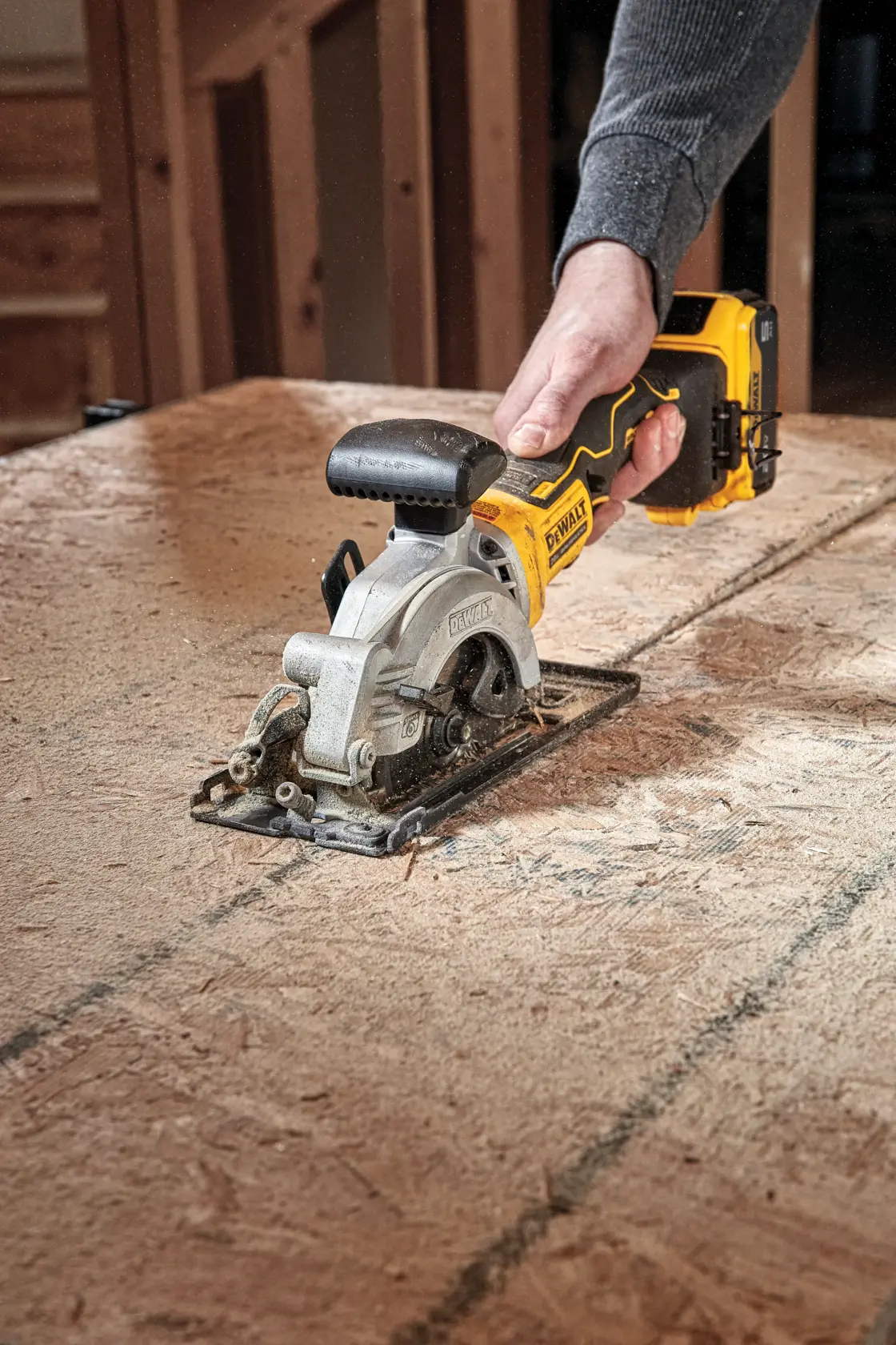 A person using a DEWALT cordless circular saw to cut a sheet of wood on a work table in a workshop.
