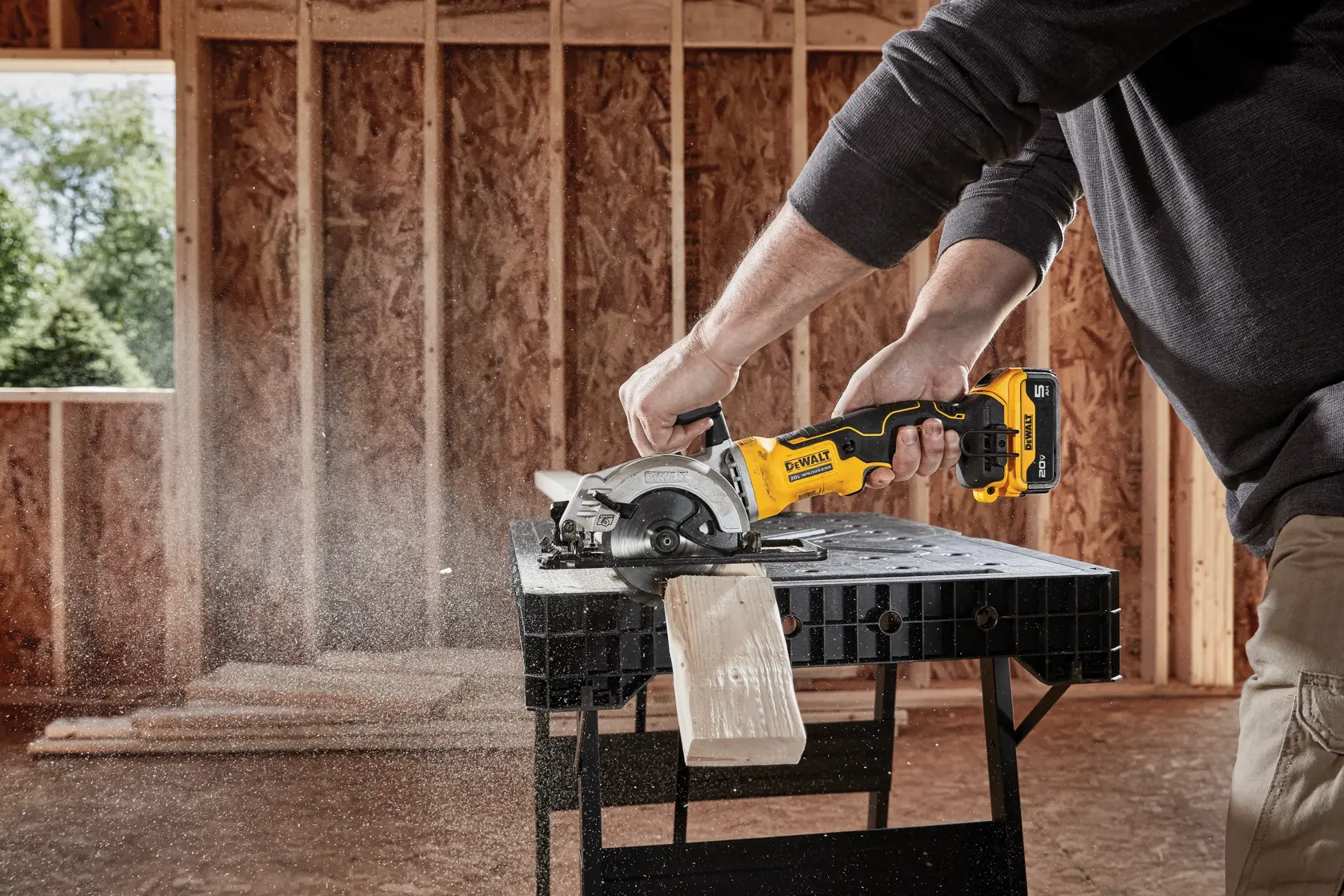 A person using a DEWALT cordless circular saw to cut a piece of wood on a workbench inside a building under construction.
