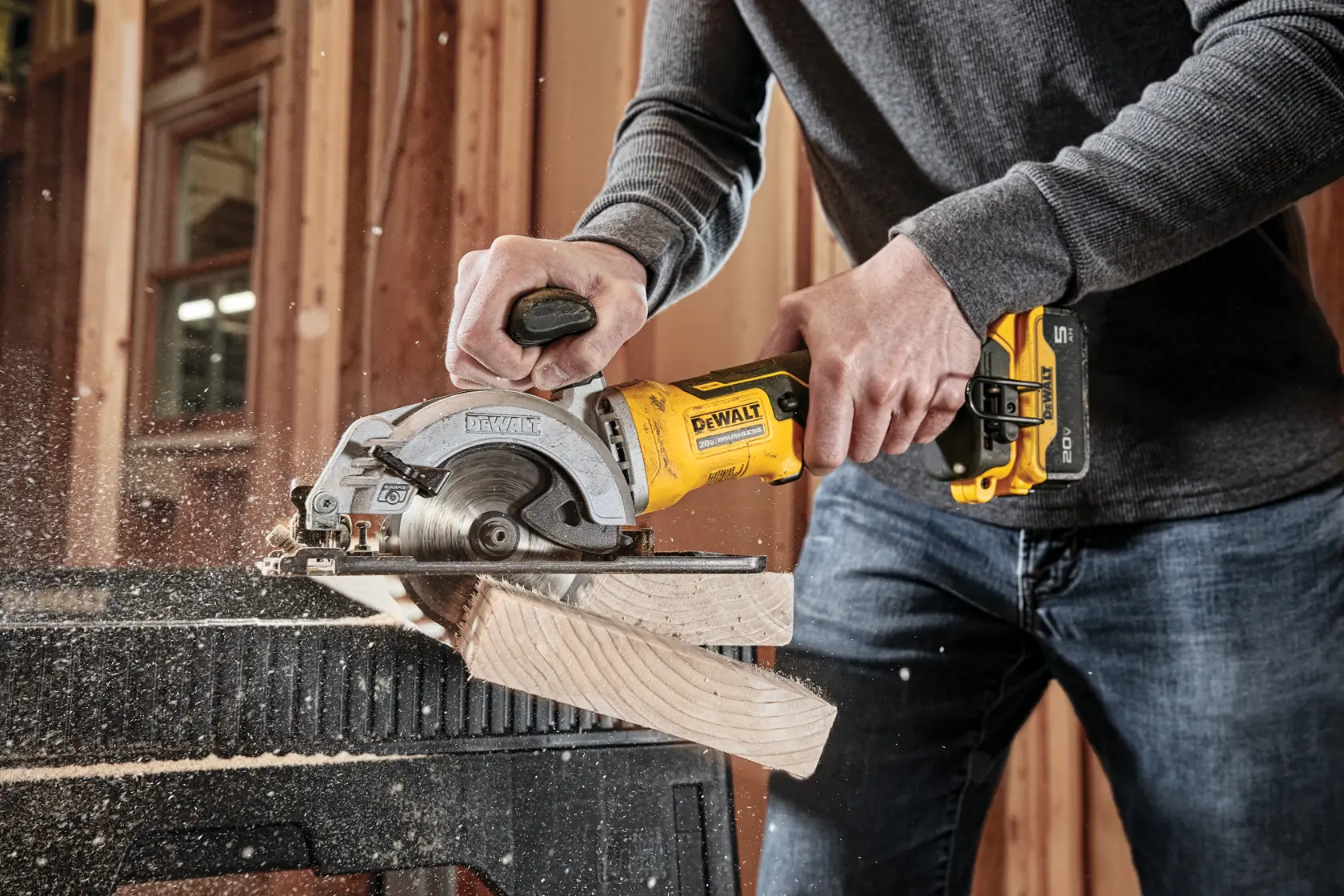 A person using a DEWALT cordless circular saw to cut a piece of wood in a workshop. Sawdust is visible as the wood is being cut.