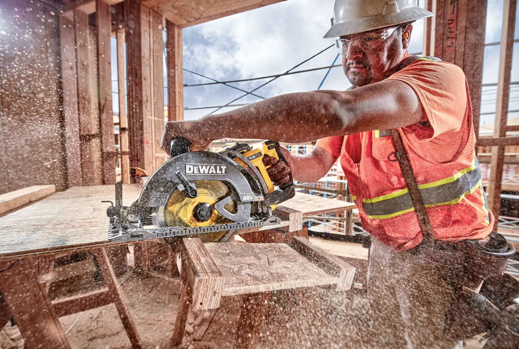 A person wearing safety gear and an orange vest uses a DEWALT circular saw (model DCS577B) to cut a wooden board at an active construction site. Sawdust is flying through the air, highlighting the power of the tool.