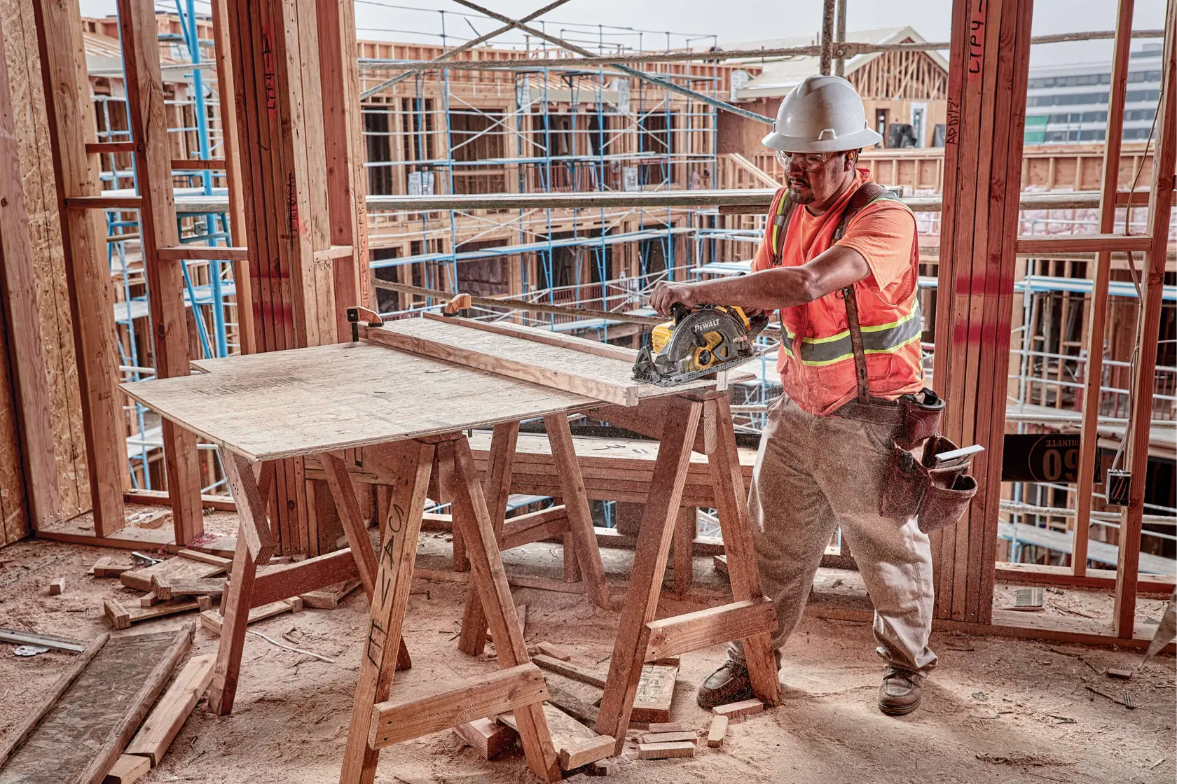 A construction worker wearing a hard hat and safety vest uses a DEWALT DCS577B circular saw to cut wood on a job site. The worker is surrounded by framing materials and scaffolding, and is working on a makeshift table made of wood.