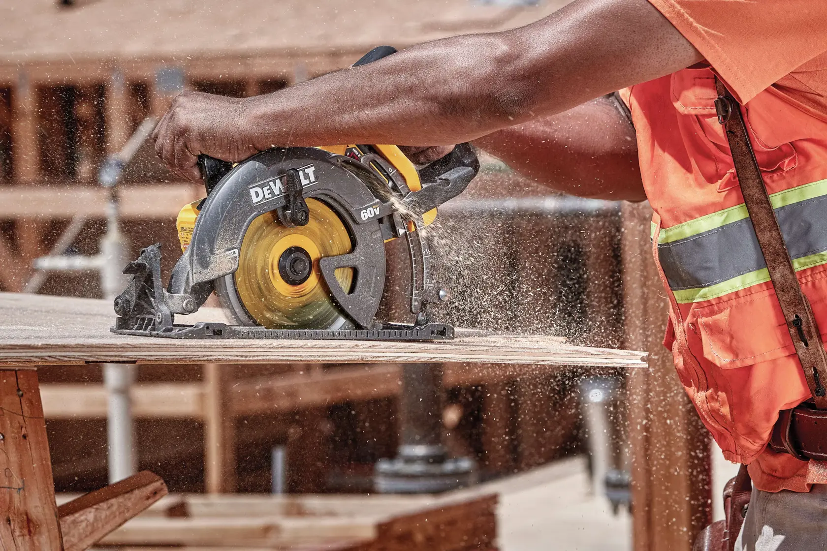 A person wearing an orange safety vest is using a DEWALT DCS577B circular saw to cut a wooden plank at a construction site. Sawdust is visible in the air.