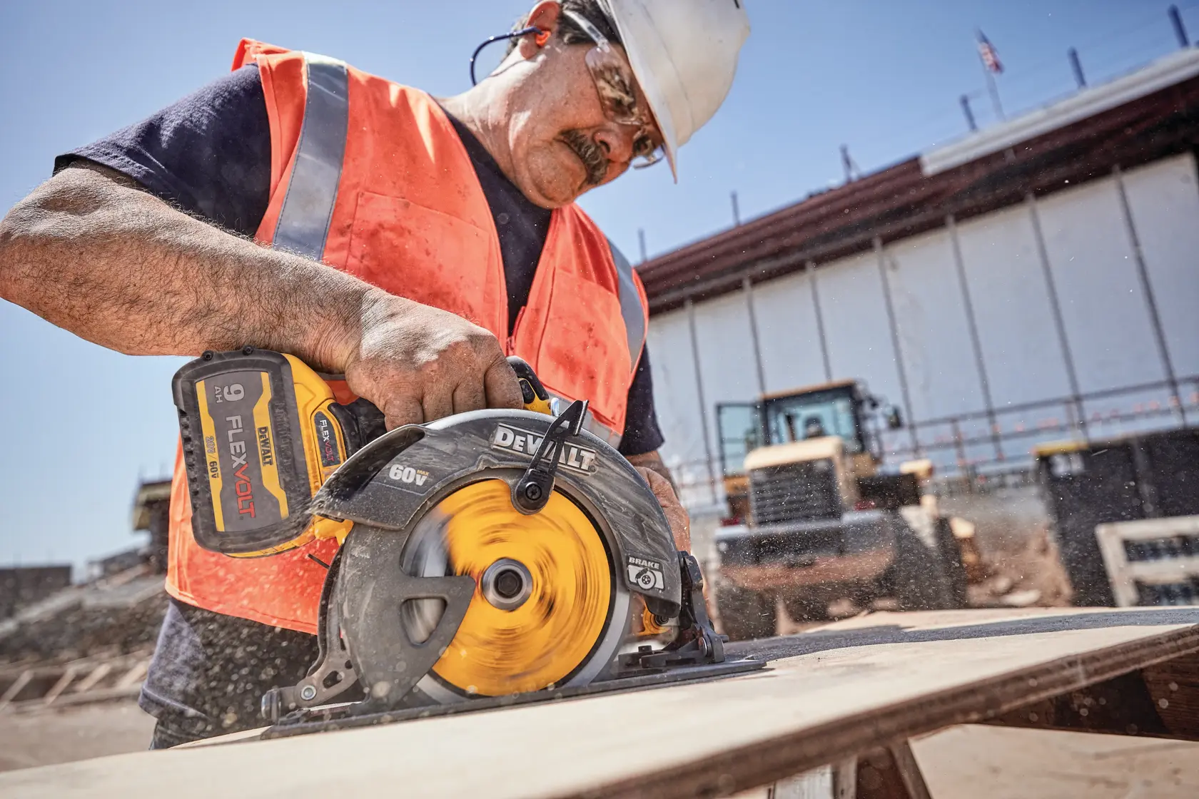 A construction worker wearing a safety vest and helmet is using a DEWALT 60V FLEXVOLT cordless circular saw to cut a wooden plank at a construction site. The worker's face is blurred for privacy.