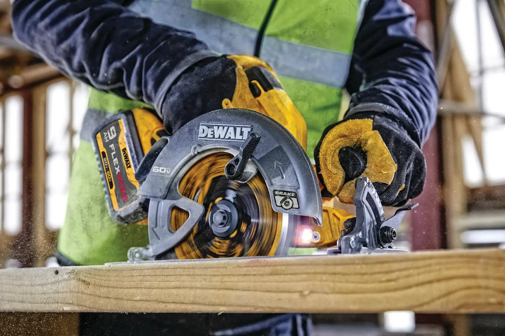 A person wearing protective gloves and a safety vest is using a DEWALT cordless circular saw to cut a piece of wood on a construction site.
