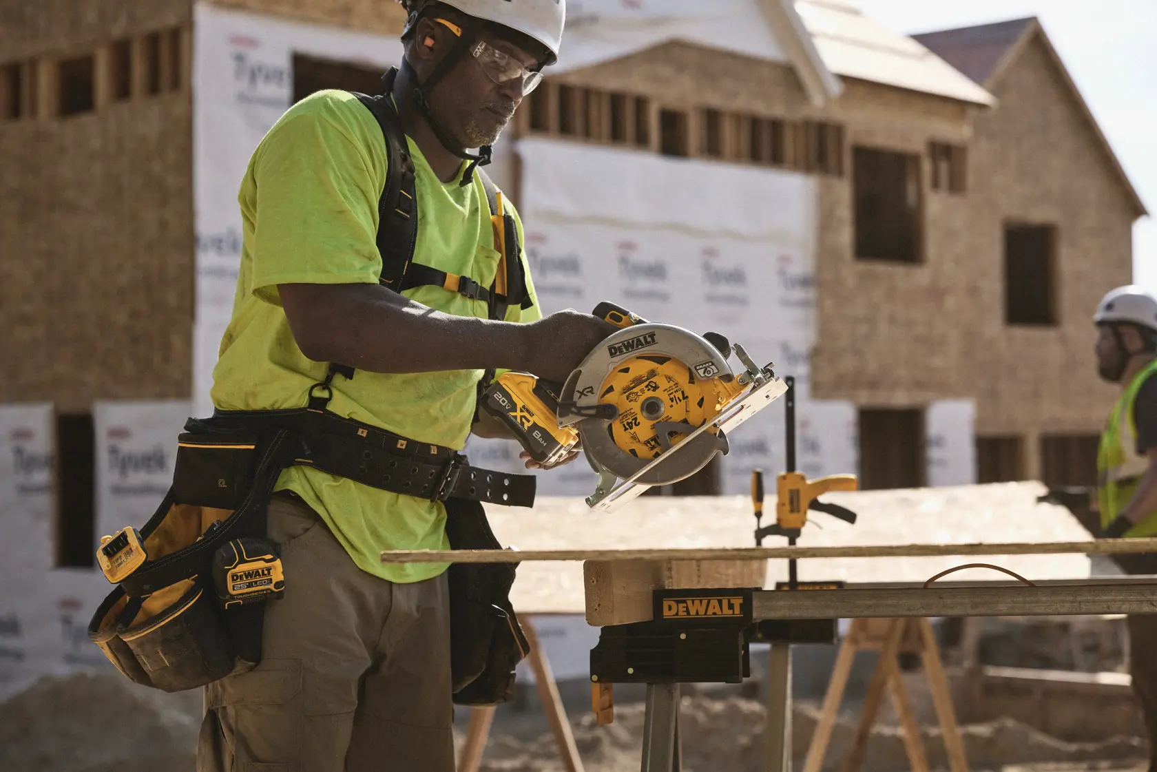A construction worker using a DEWALT circular saw to cut wood at a building site. The worker is wearing a tool belt and safety gear, and there are unfinished houses in the background.