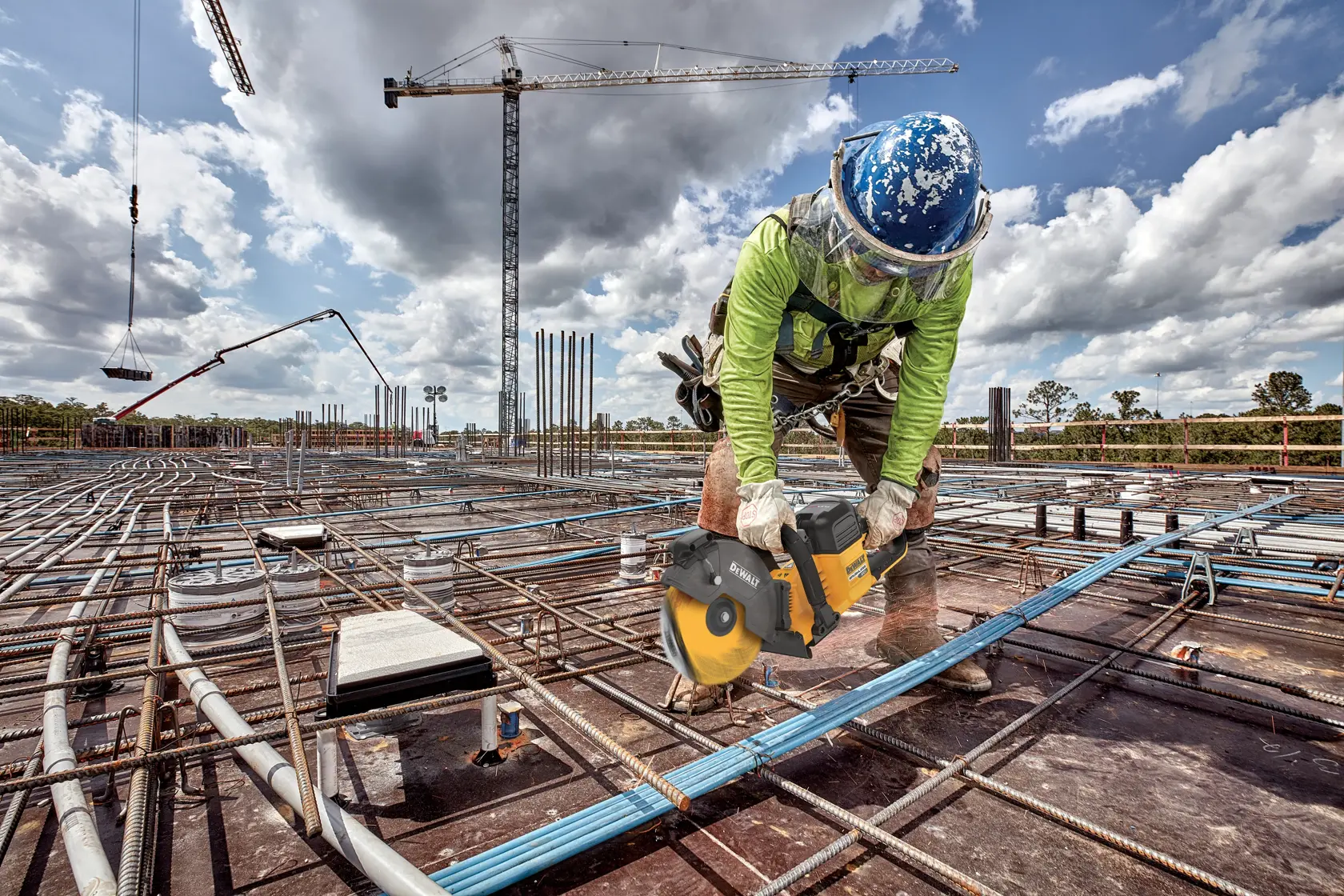 A construction worker wearing safety gear is using a DEWALT cordless saw to cut through a pipe or conduit on a building site filled with steel reinforcement bars and construction equipment. Cranes and a cloudy sky are visible in the background.
