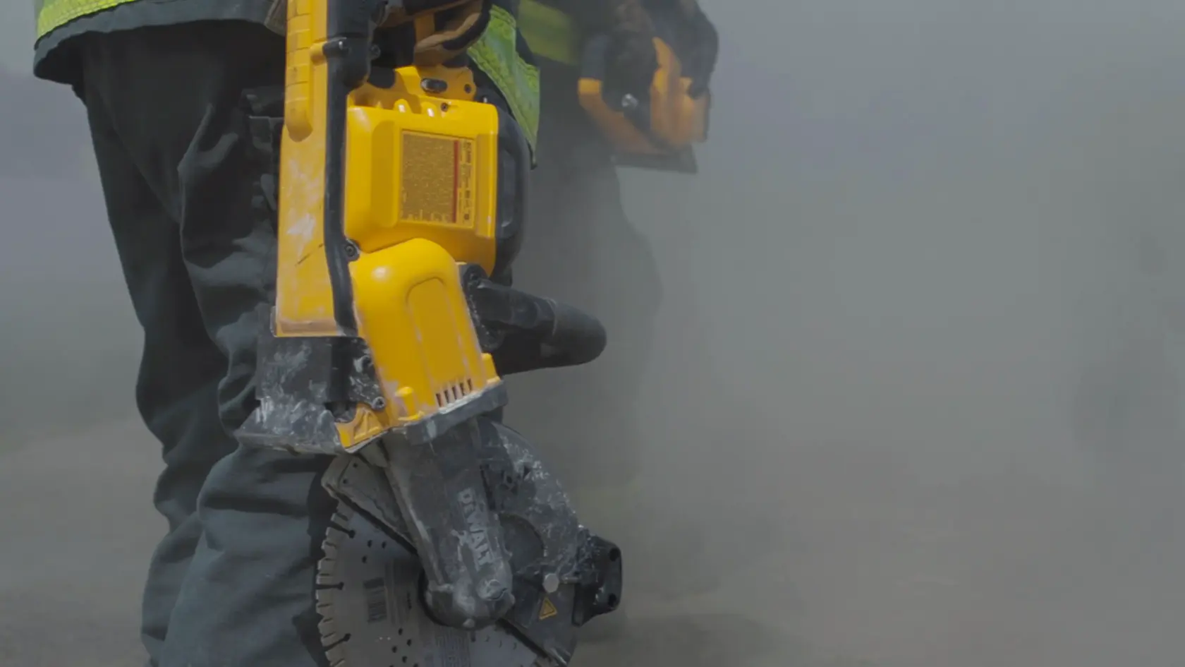 Close-up of a person in work clothes operating a DEWALT yellow power tool, likely a saw, amidst dust on a construction site.