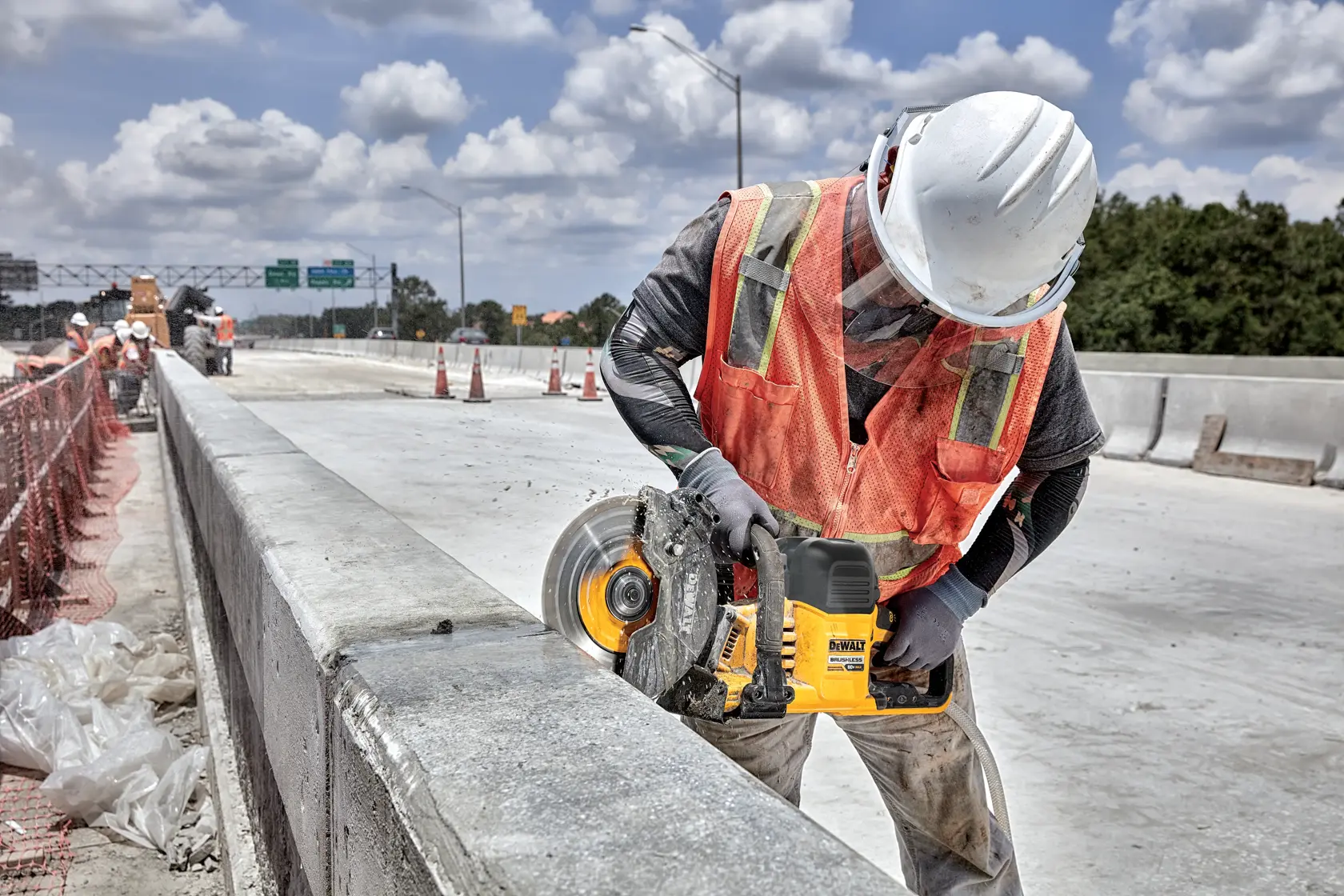A construction worker wearing a white hard hat and orange safety vest is using a DEWALT DCS692X2 power saw to cut concrete on a roadway construction site.