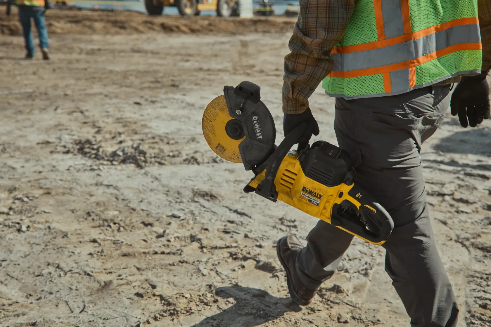 A person wearing a high-visibility vest and gloves is carrying a DEWALT DCS692X2 cordless cut-off saw at a construction site.