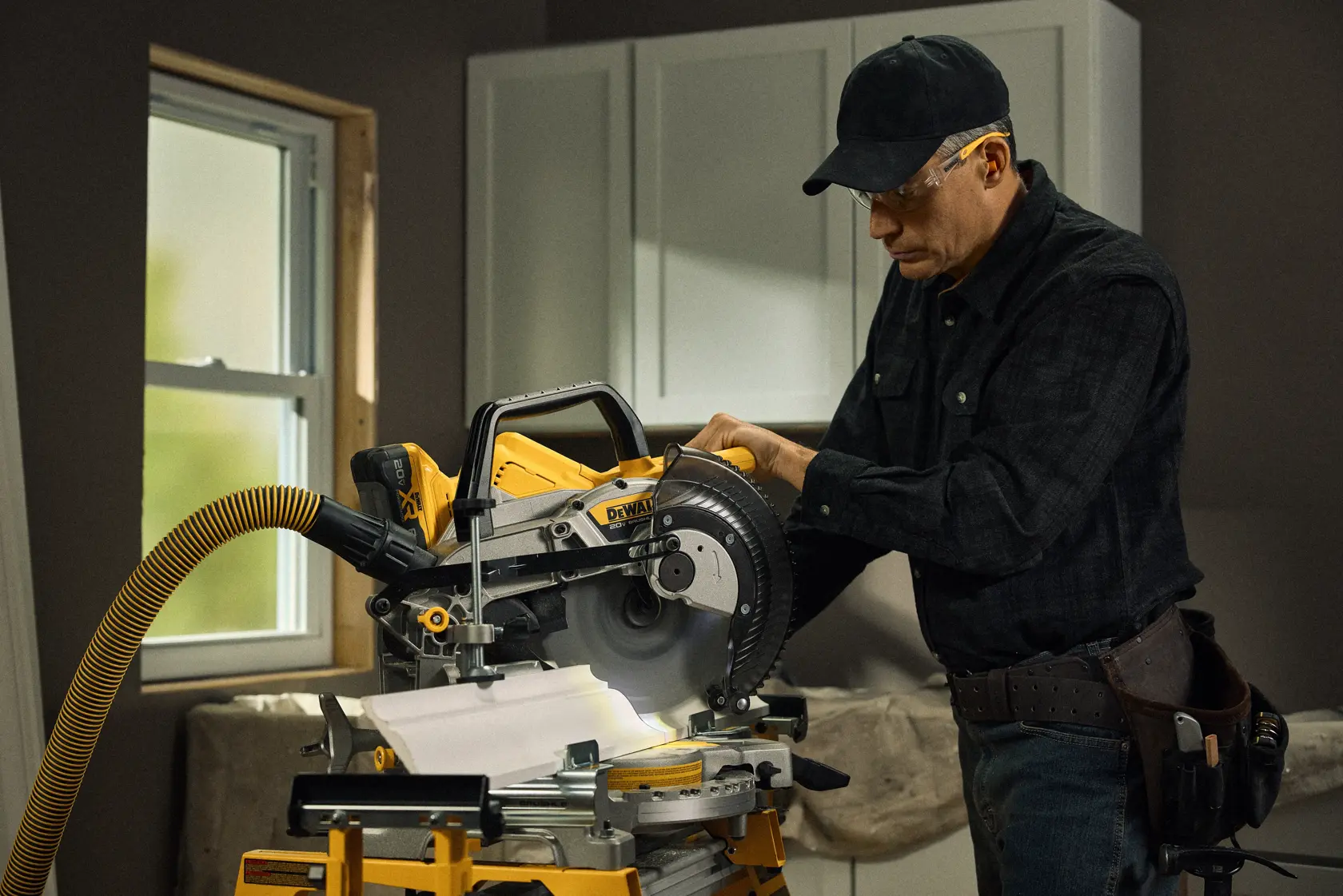 A person using a DEWALT DCS714B miter saw in a workshop setting, cutting a piece of wood. The saw is connected to a yellow vacuum hose for dust collection. The workshop has cabinets and a window in the background.