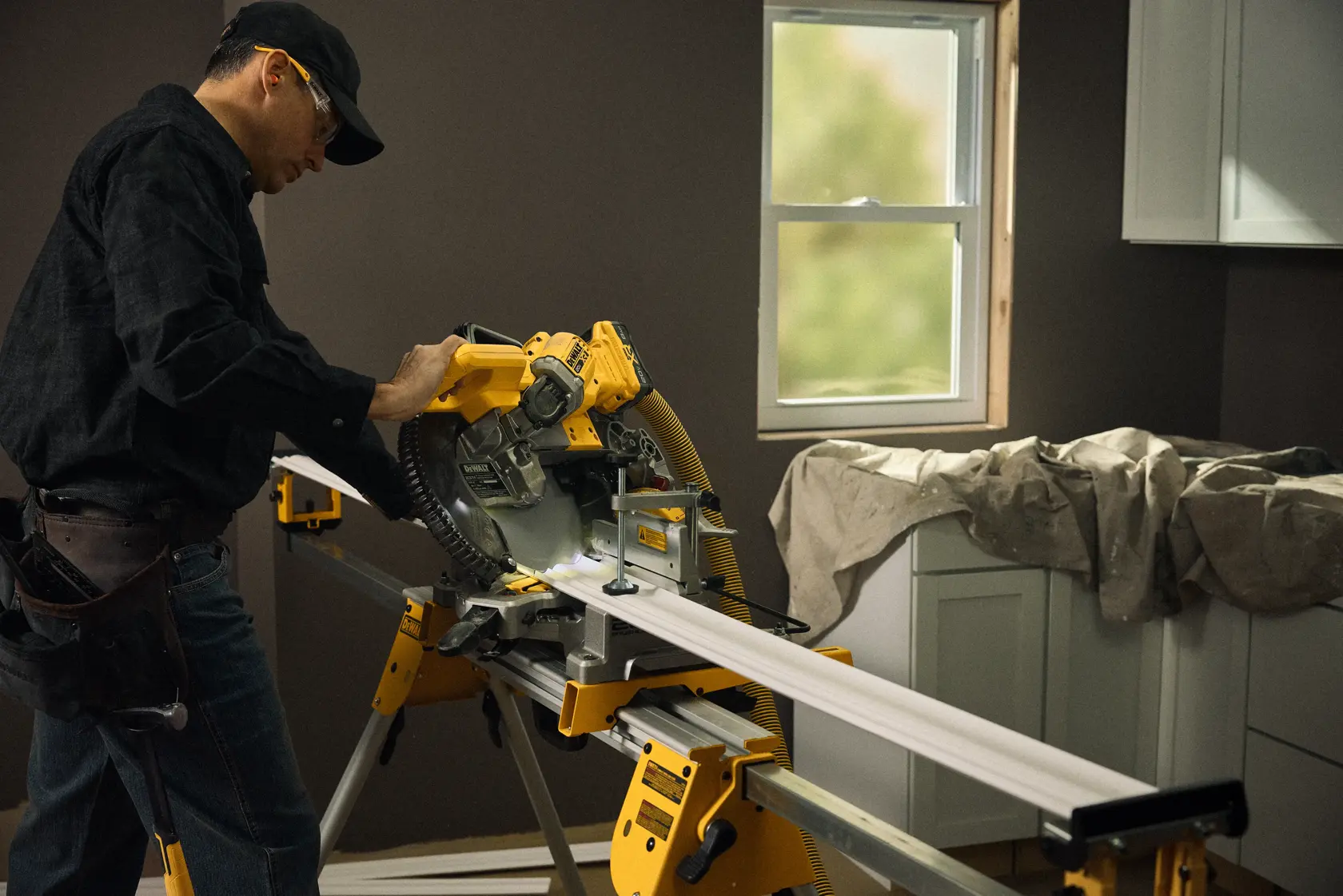 A person using a yellow DEWALT miter saw to cut a piece of white molding inside a room. The saw is placed on a work stand, and the person is wearing safety glasses and a tool belt. The background shows a window and covered cabinets.