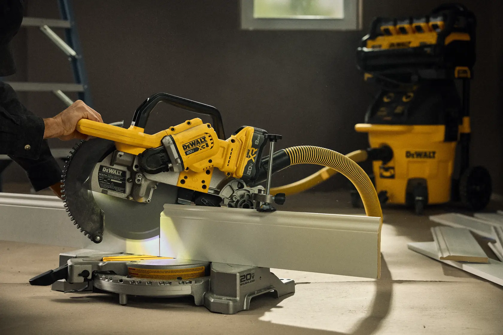 A person is using a DEWALT cordless miter saw with a yellow vacuum hose attached, cutting a piece of white trim inside a workshop. There is a DEWALT dust extractor in the background, along with other pieces of trim and a ladder.