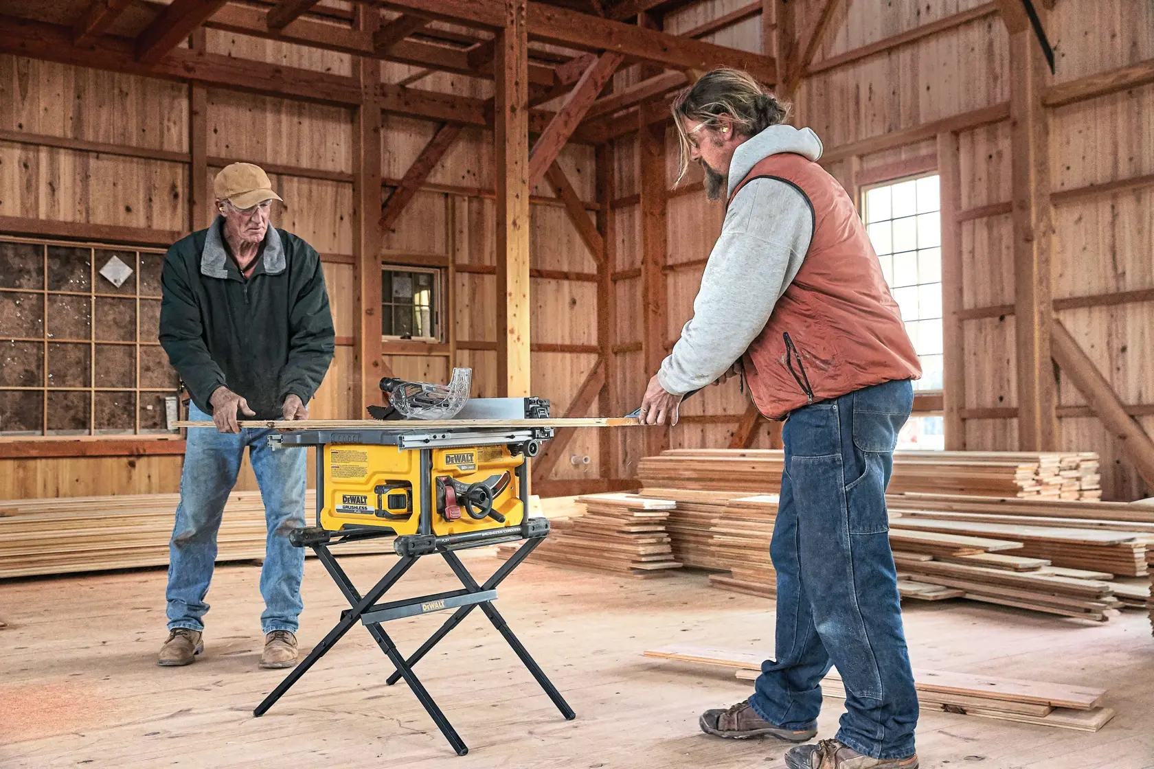Two people are working together in a wooden workshop, using a yellow DEWALT table saw placed on a stand to cut a piece of wood. The background shows stacks of wood and wooden walls, with natural light coming through the windows.
