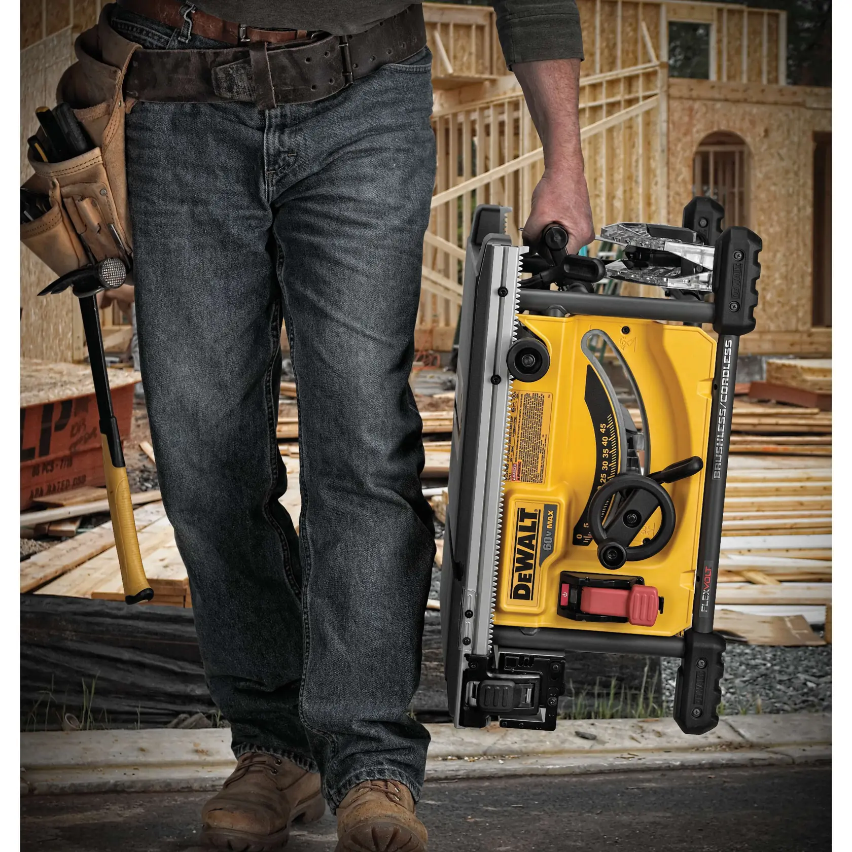A person wearing jeans and work boots carries a portable DEWALT table saw at a construction site. The worker is also holding a hammer and wearing a tool belt. The background shows unfinished wooden structures.