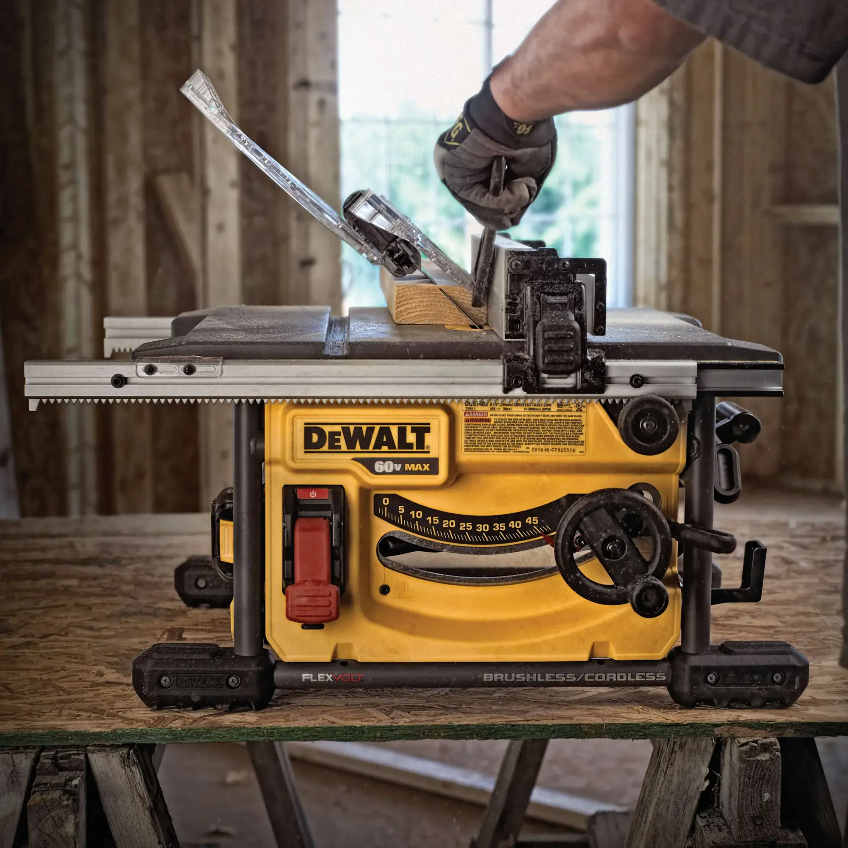 A DEWALT cordless table saw positioned on a wooden workbench in a construction setting. A person wearing a glove is adjusting a piece of wood for cutting. The saw features clear safety guards and visible adjustment controls.