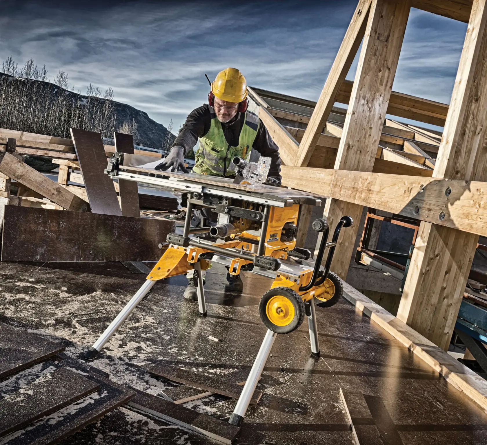 A DEWALT portable table saw with yellow accents is set up outdoors at a construction site. A person wearing a hard hat, safety vest, ear protection, and gloves is operating the saw, surrounded by wooden beams and boards, with mountains and trees visible in the background.