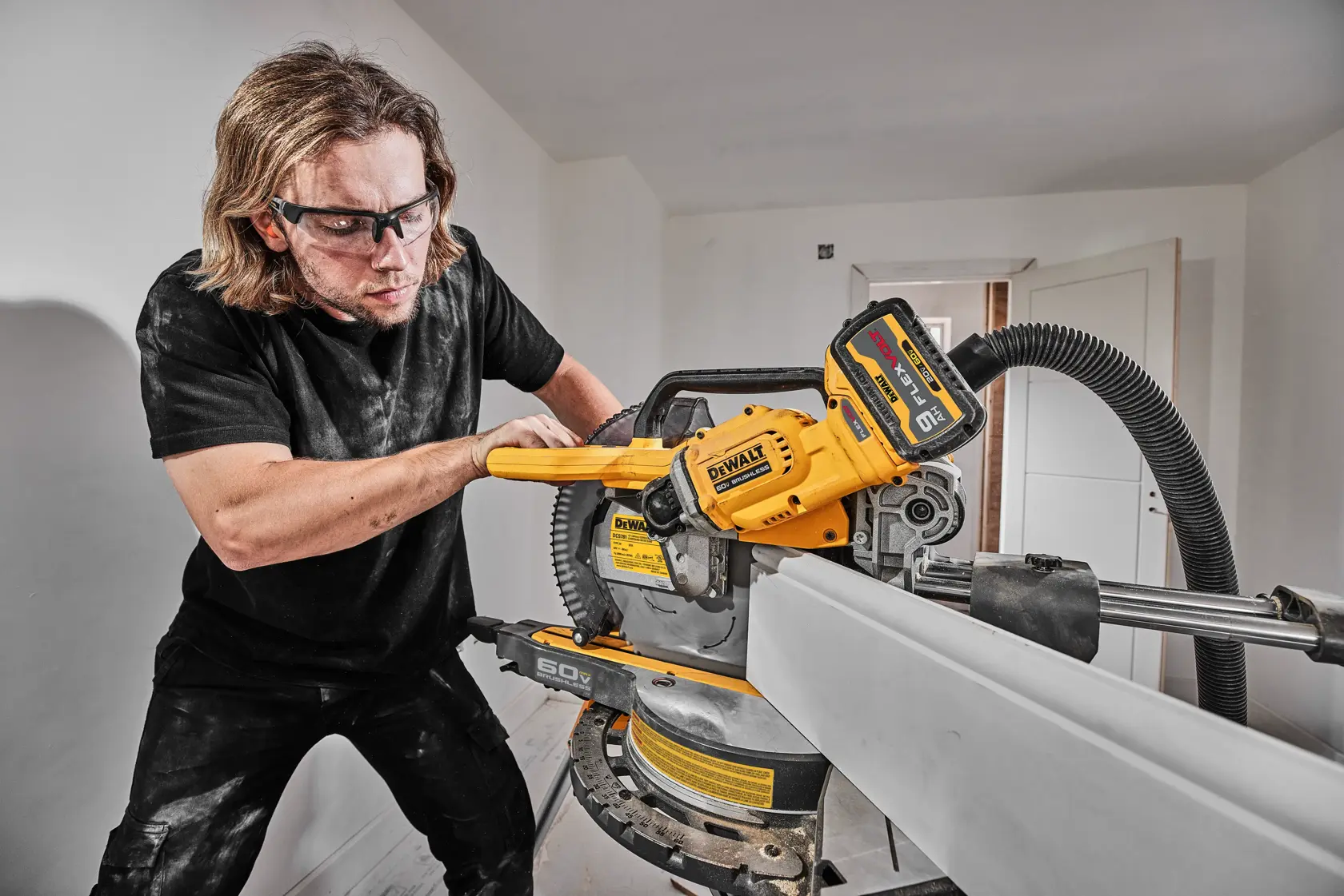 A person operates a DEWALT cordless miter saw (model DCS781B) indoors, making a cut on a long piece of material. The tool is connected to a dust extraction hose. The face of the person is blurred for privacy.