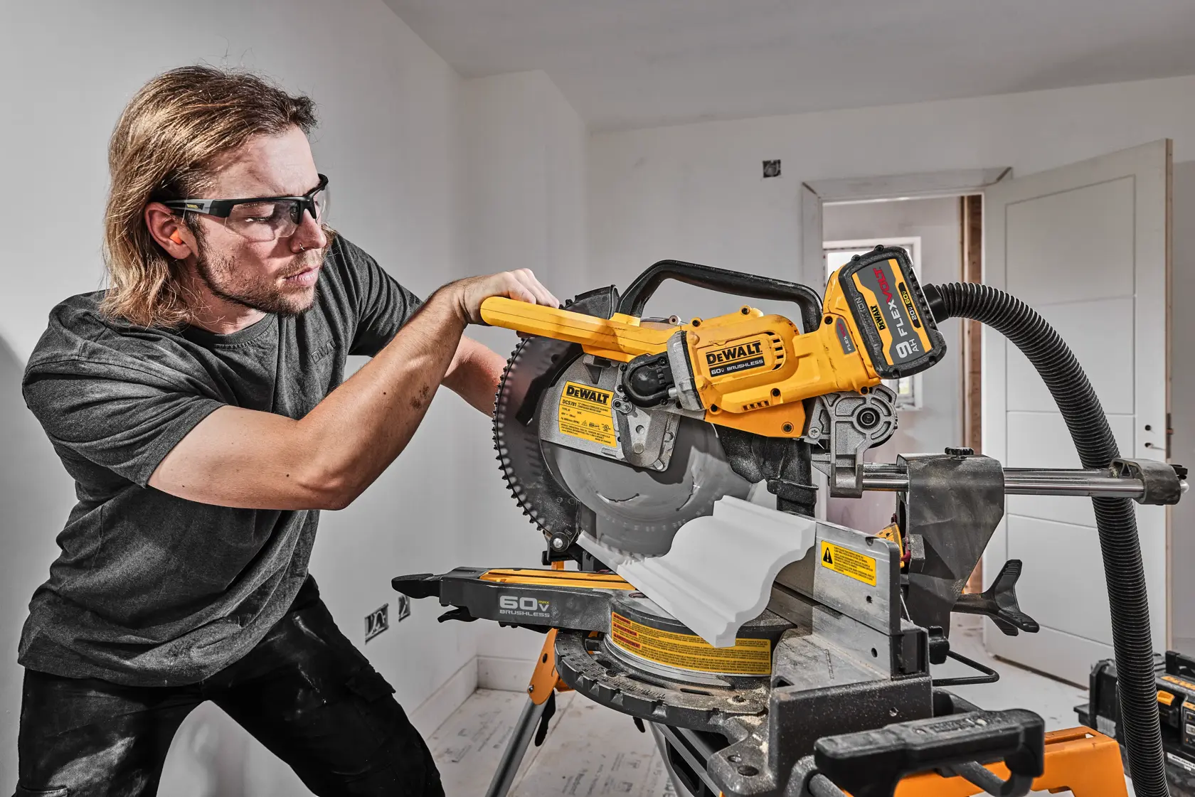 A person using a DEWALT DCS781X1 miter saw to cut a piece of white molding in a well-lit indoor setting. The saw is yellow and black with safety labels and a hose attached for dust extraction. The person's face is blurred for privacy.