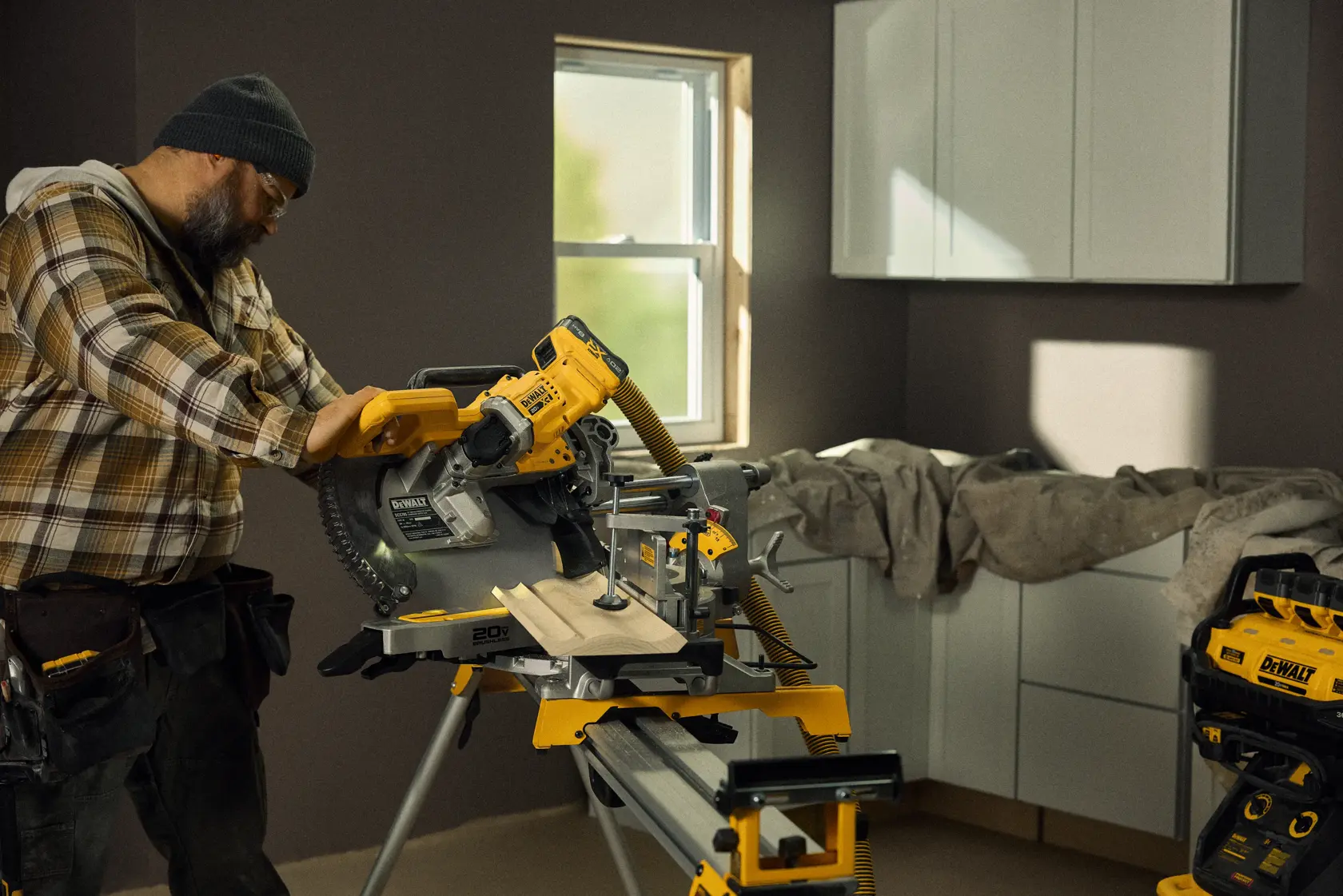 A person operating a DEWALT cordless miter saw (model DCS785B) to cut wooden planks inside a workshop or renovated room. The background features cabinets, a window, and other DEWALT tools and equipment.
