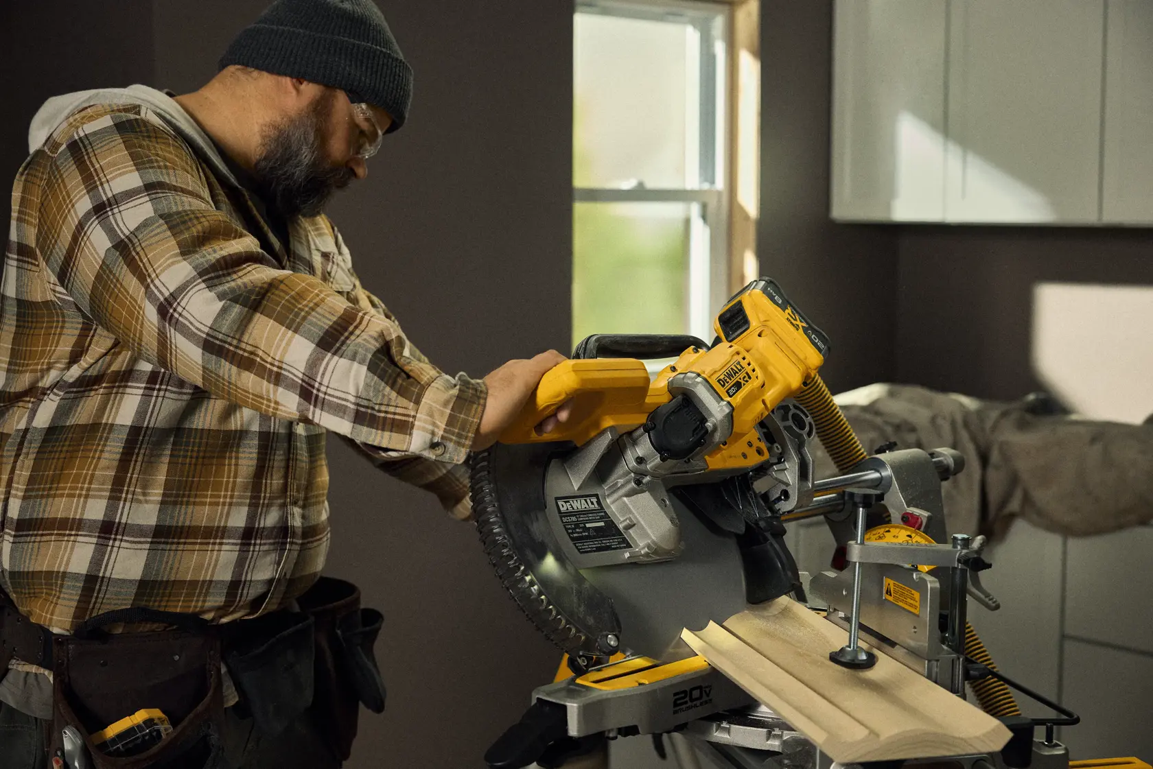 A person operating a DEWALT DCS785B cordless miter saw to cut wooden trim in a well-lit indoor workshop. The worker is wearing a plaid shirt, a tool belt, and a beanie. The saw is positioned on a work bench, and the room has cabinetry and a window.