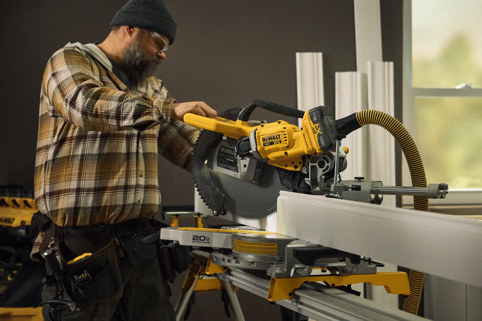 A person in a plaid shirt and beanie using a DEWALT DCS785WW1 cordless miter saw to cut a long piece of white material in an indoor workshop setting. The saw is attached to a yellow and gray stand, and there is a flexible vacuum hose connected to the tool for dust extraction.