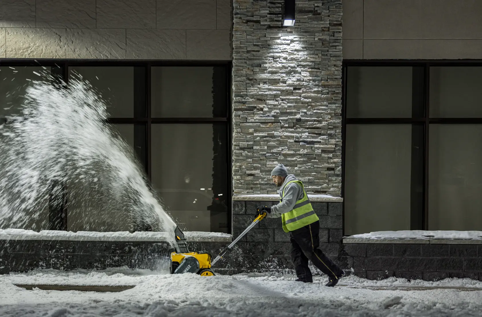A person wearing a reflective vest uses a DEWALT DCSNP2142 snow blower to clear snow from a sidewalk at night, with snow being thrown to the side.