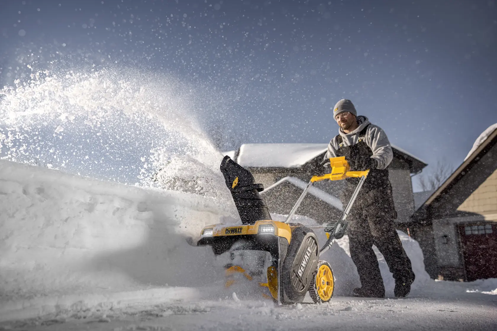 A person using a DEWALT snow blower to clear snow outside a house, with snow being discharged from the machine.