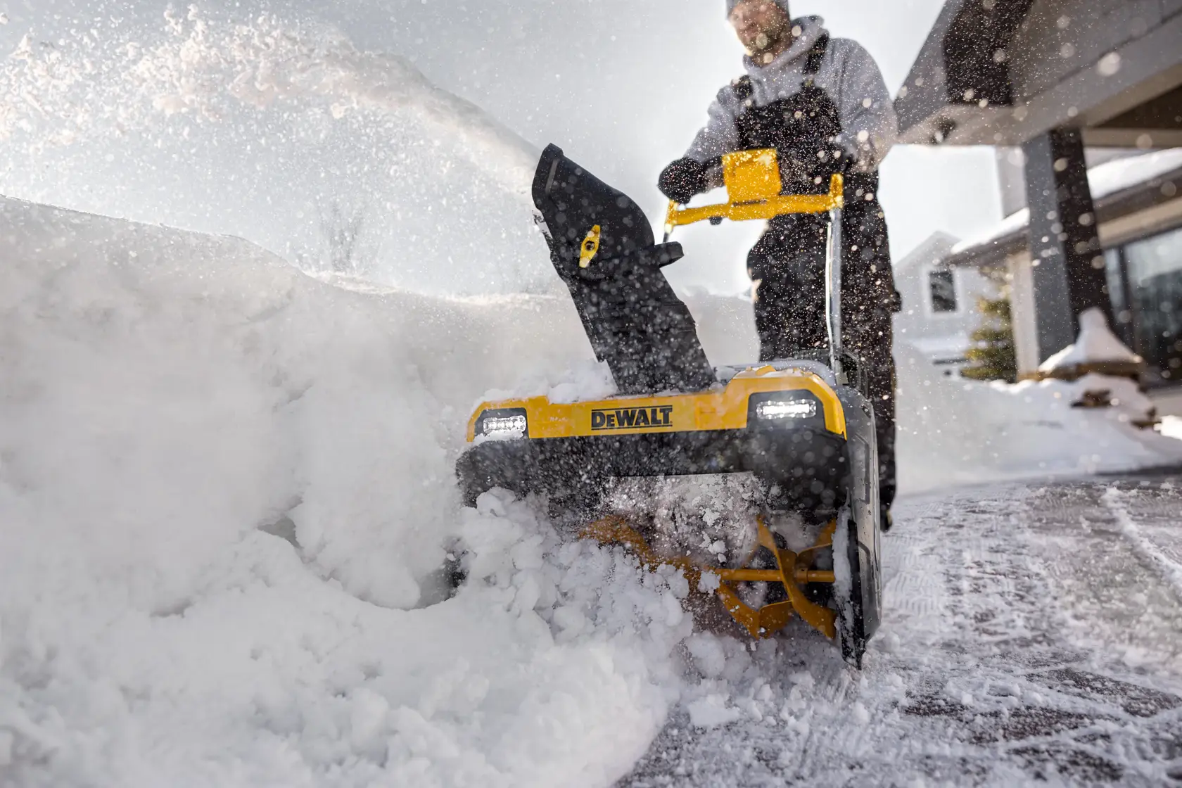 A person uses a DEWALT snow blower to clear snow from a driveway in front of a house. The snow blower shoots snow to the side as the person pushes it forward.