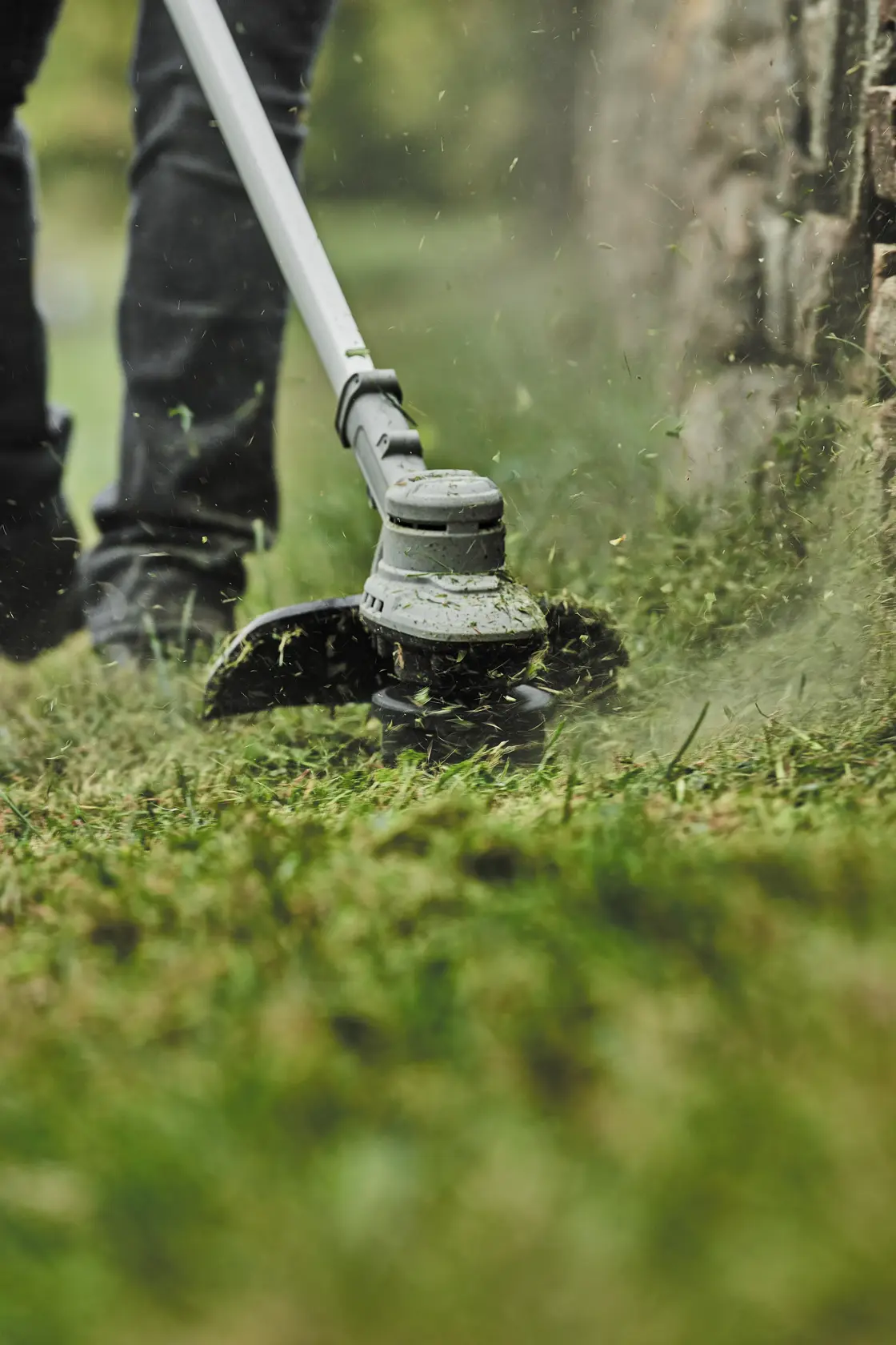 Close-up of a person using a DEWALT string trimmer to cut grass near a stone wall.