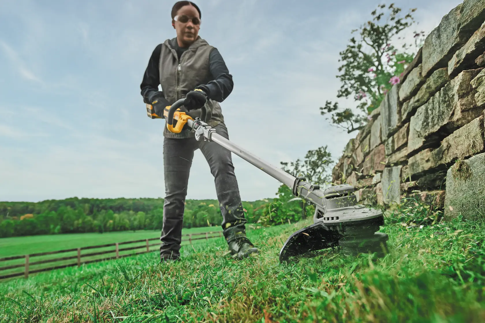 A person using a DEWALT cordless string trimmer to cut grass along a stone wall outdoors, with a green landscape and blue sky in the background. The person's face is blurred.
