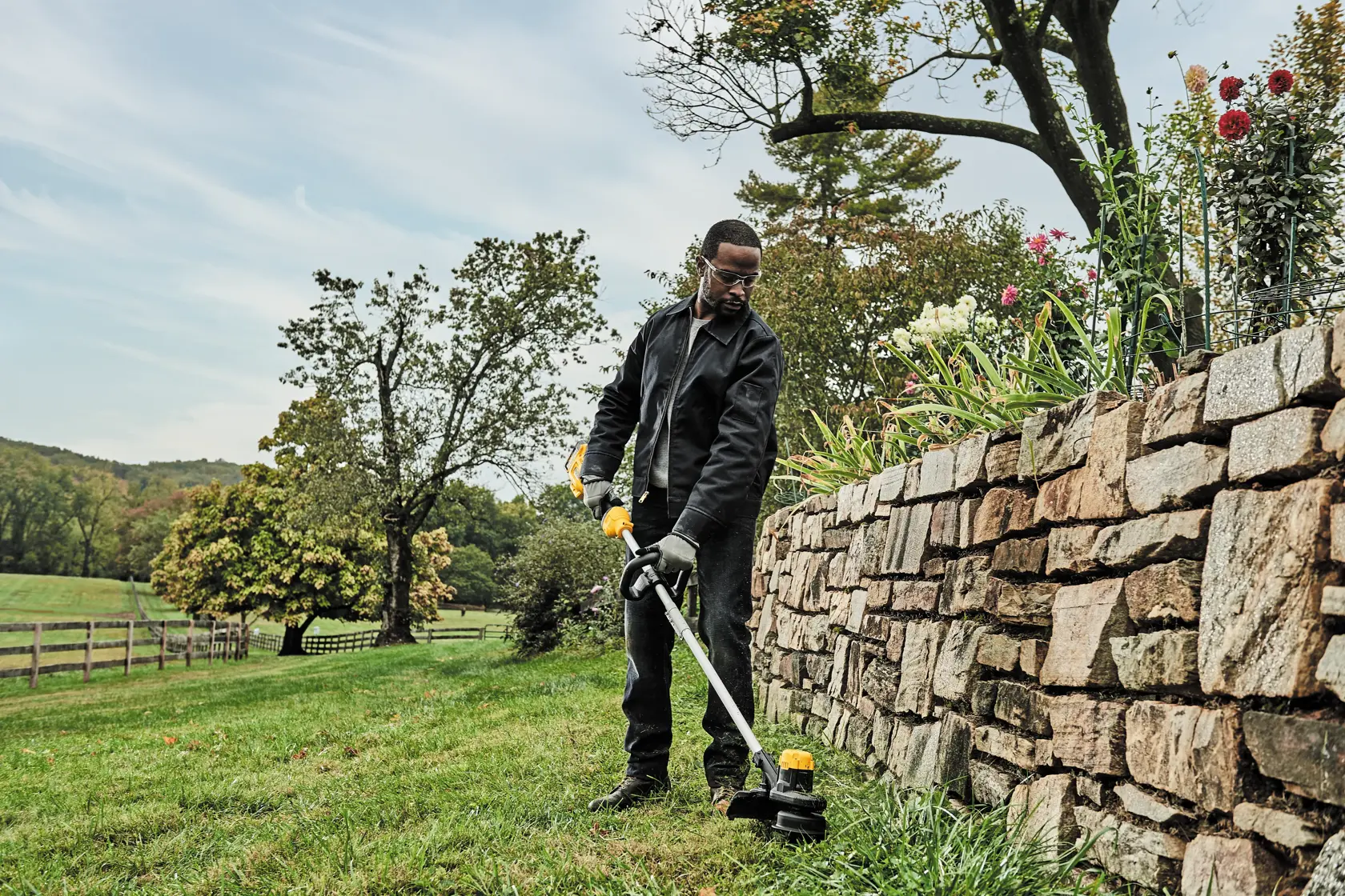 A person using a DEWALT cordless string trimmer (model DCST925B) to trim grass along a stone wall in a garden or outdoor area. The individual is wearing protective gloves and dark clothing. The landscape features green grass, trees, and flowers growing above the wall.