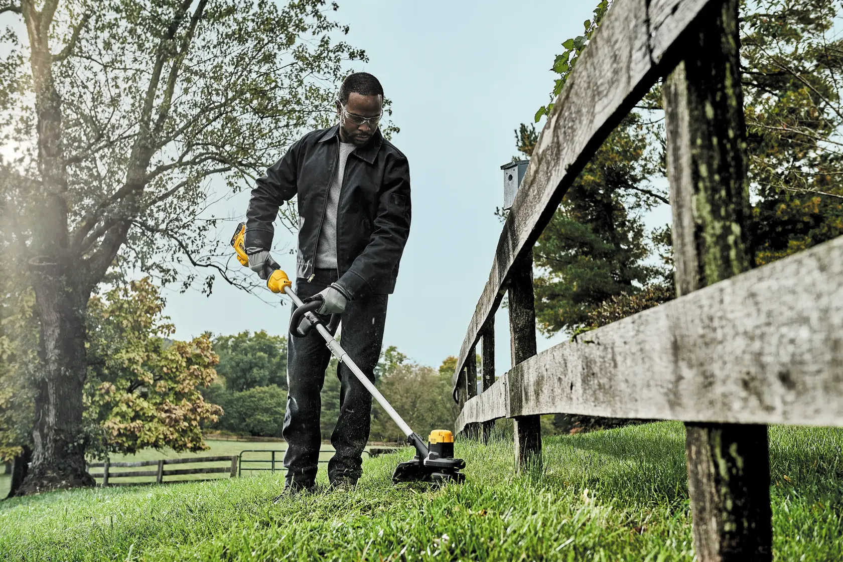 A person using a DEWALT DCST925B cordless string trimmer to cut grass near a wooden fence in an outdoor setting, with trees and greenery in the background.