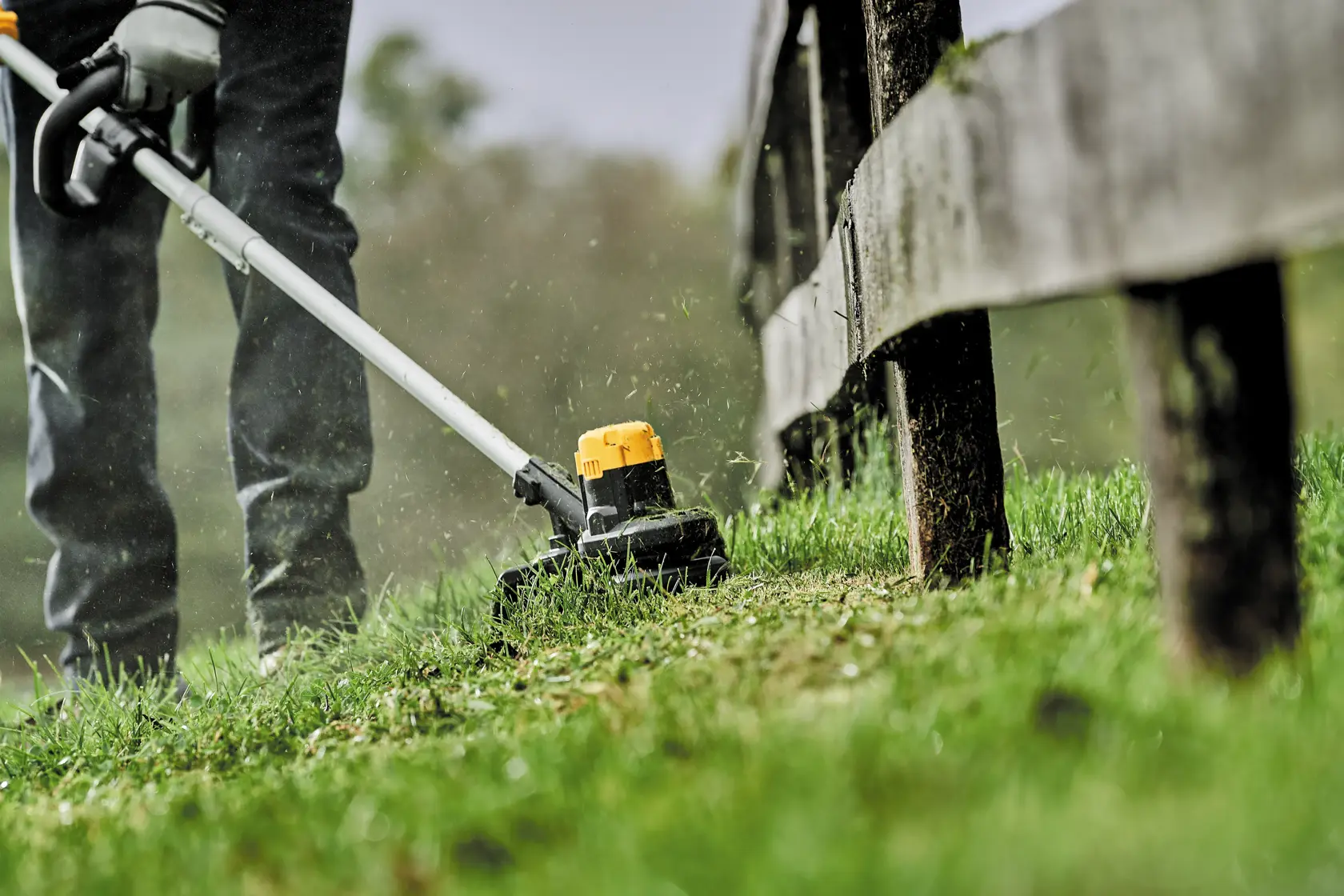 A person is using a DEWALT cordless string trimmer to trim grass along a wooden fence outdoors.