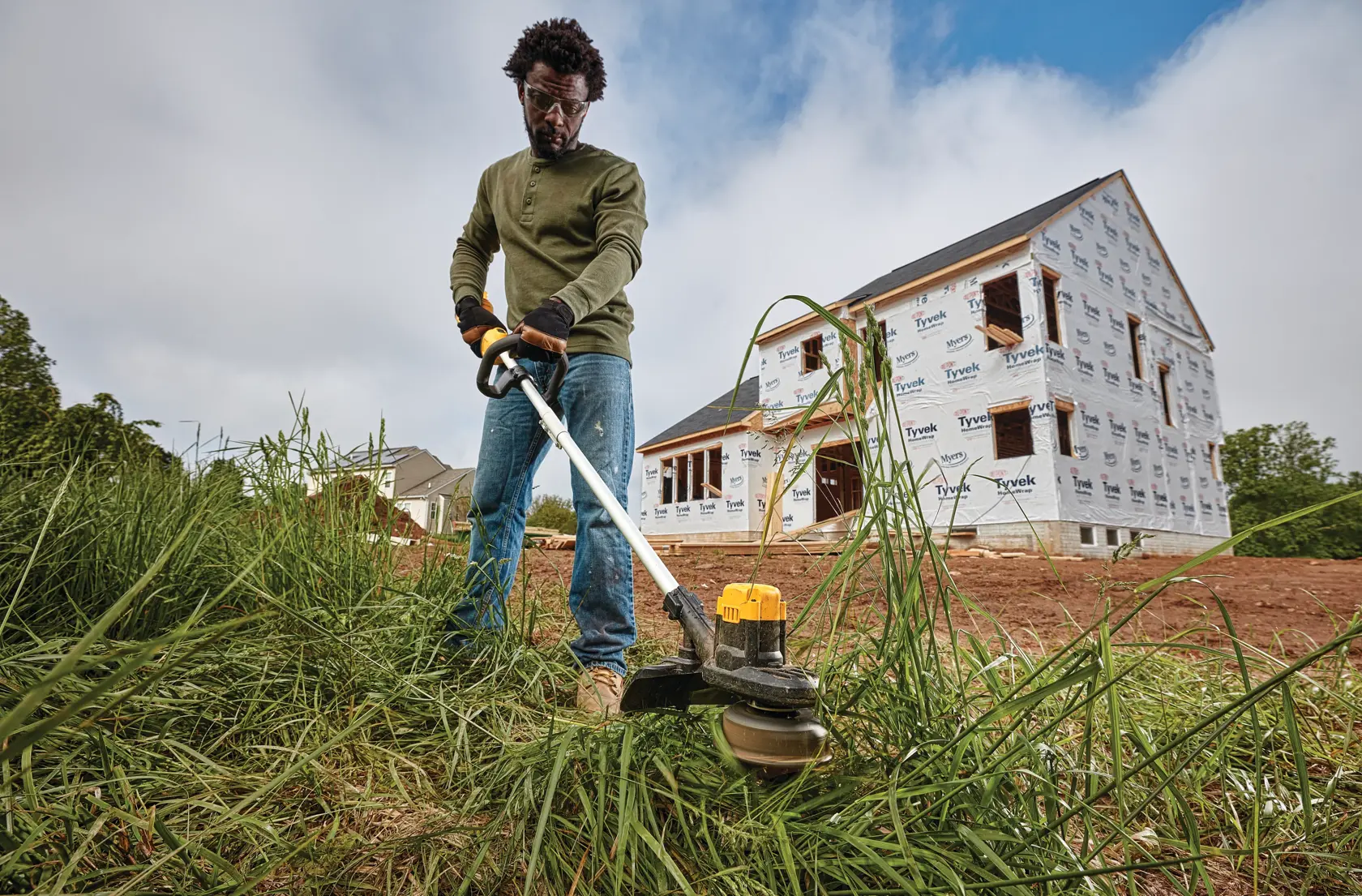 A person using a DEWALT cordless string trimmer to cut tall grass in front of a house under construction. The person is standing outdoors, and the house has Tyvek wrapping visible on the exterior.