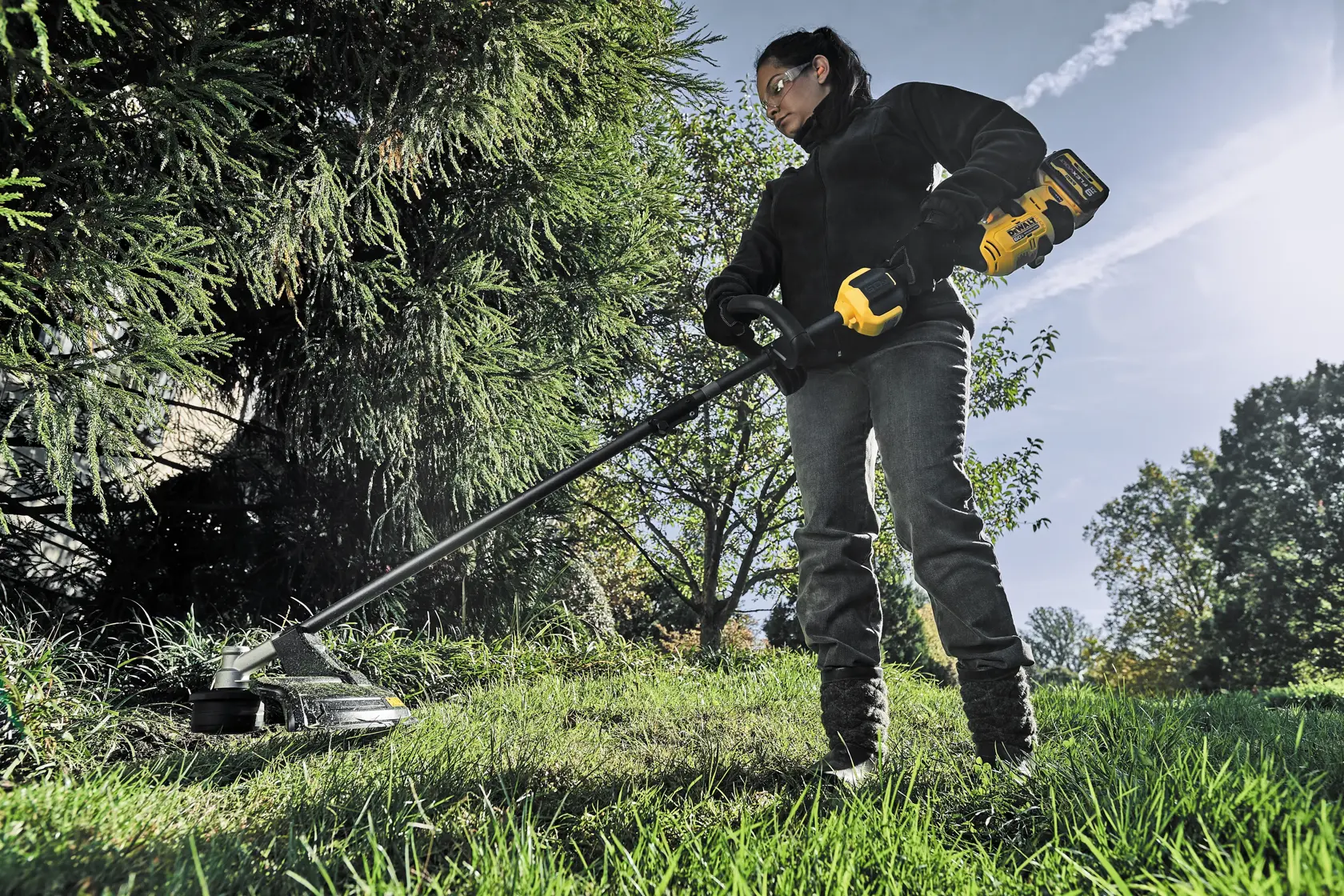 A person outdoors is operating a DEWALT DCST972X1 cordless string trimmer, trimming grass near bushes and trees.