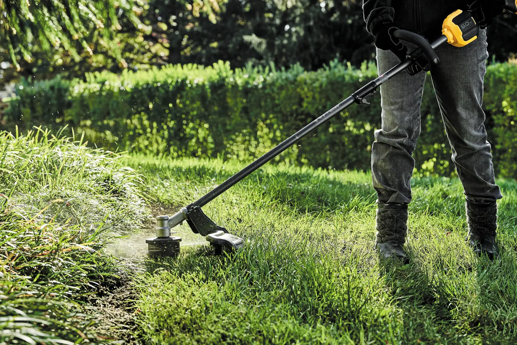A person uses a DEWALT cordless string trimmer to cut grass along the edge of a lawn on a sunny day.