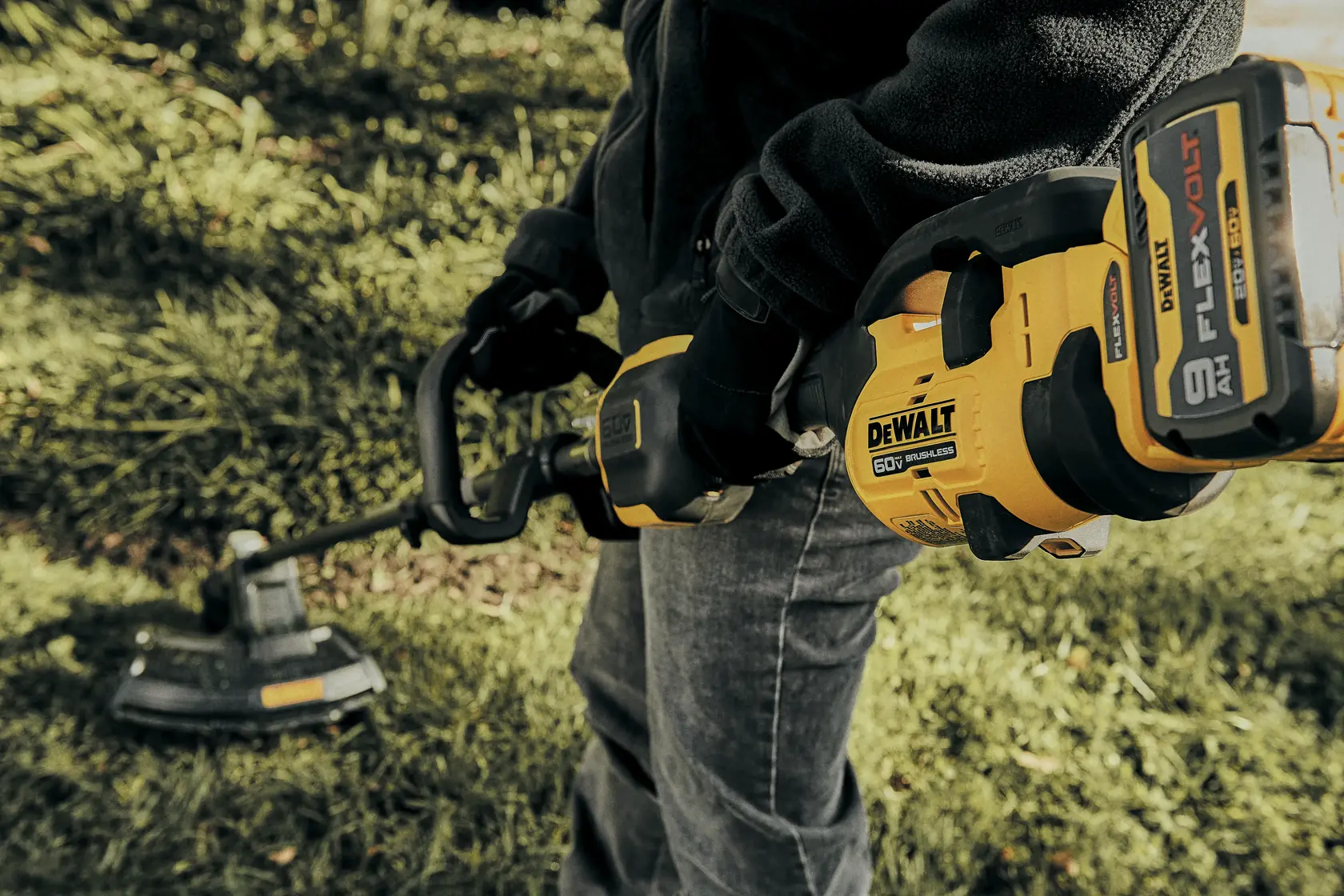 A person holding a DEWALT DCST972X1 battery-powered string trimmer with a FLEXVOLT 60V lithium-ion battery, being used outdoors on grass.