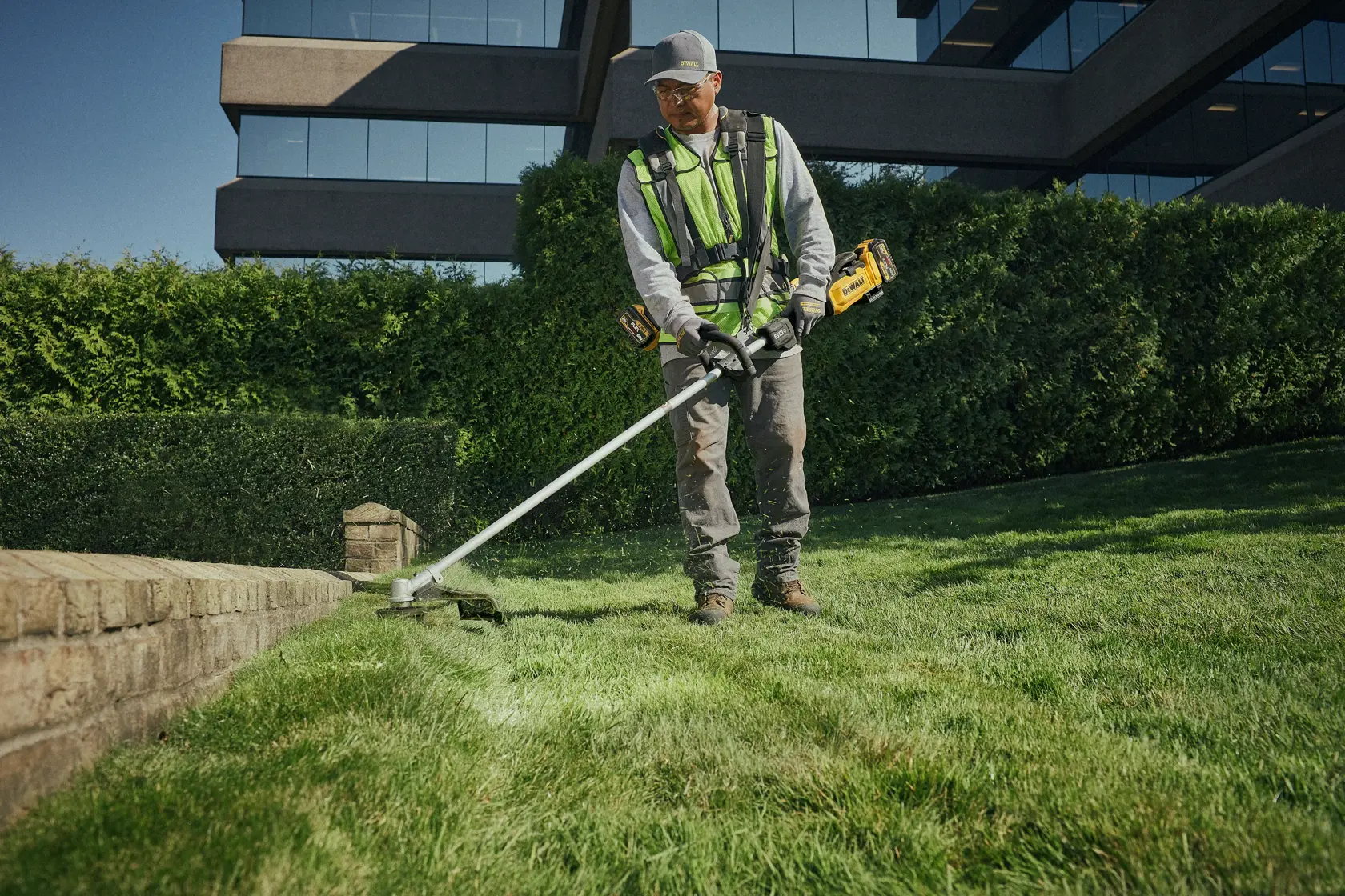 A person wearing a safety vest and gloves is using a DEWALT cordless string trimmer to edge the grass along a stone border in front of a modern building surrounded by hedges.