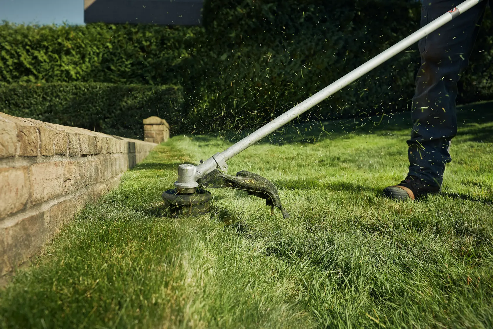 A DEWALT string trimmer being used to trim grass along a stone border in a yard. Grass clippings are visible flying from the trimmer head.