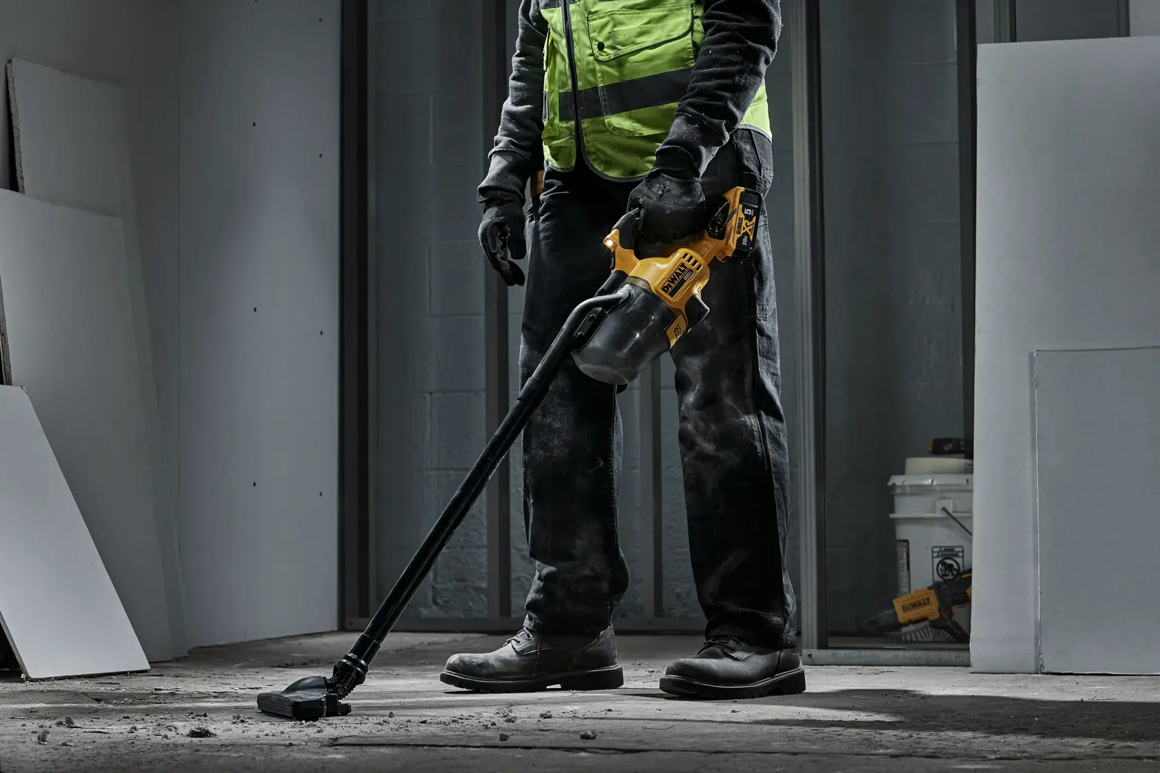 A person wearing a high-visibility safety vest and gloves is using a yellow and black DEWALT cordless vacuum cleaner to clean dust and debris from a concrete floor in an unfinished room.