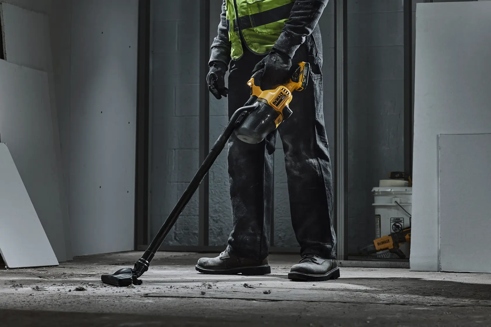 A person wearing work gloves and boots uses a DEWALT handheld vacuum cleaner with a long attachment to clean debris on a construction site floor.