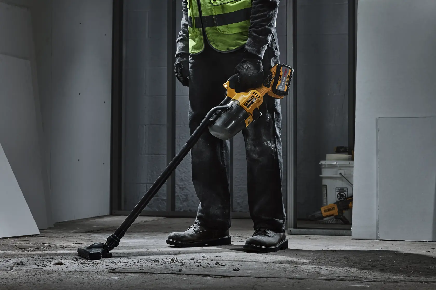 A person wearing work gloves and boots uses a DEWALT cordless handheld vacuum with a long nozzle to clean debris from the floor in a construction or renovation setting.