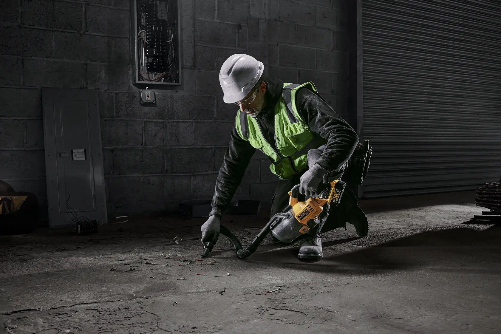 A worker wearing a safety vest and hard hat is kneeling and using a yellow DEWALT cordless vacuum to clean debris from a concrete floor in an industrial setting.