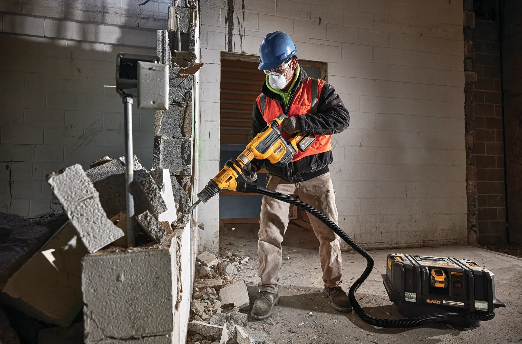 A construction worker wearing a blue hard hat and orange safety vest operates a DEWALT DCV585B dust extractor connected to a demolition tool, breaking concrete blocks inside a building.