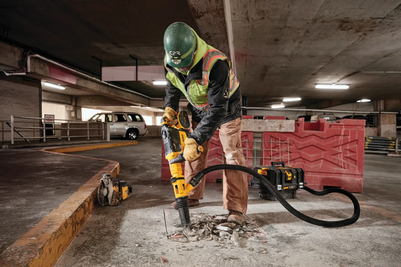 A construction worker wearing a helmet, safety vest, and gloves uses a yellow DEWALT demolition tool connected to a vacuum system to break concrete on the floor of an indoor parking garage.