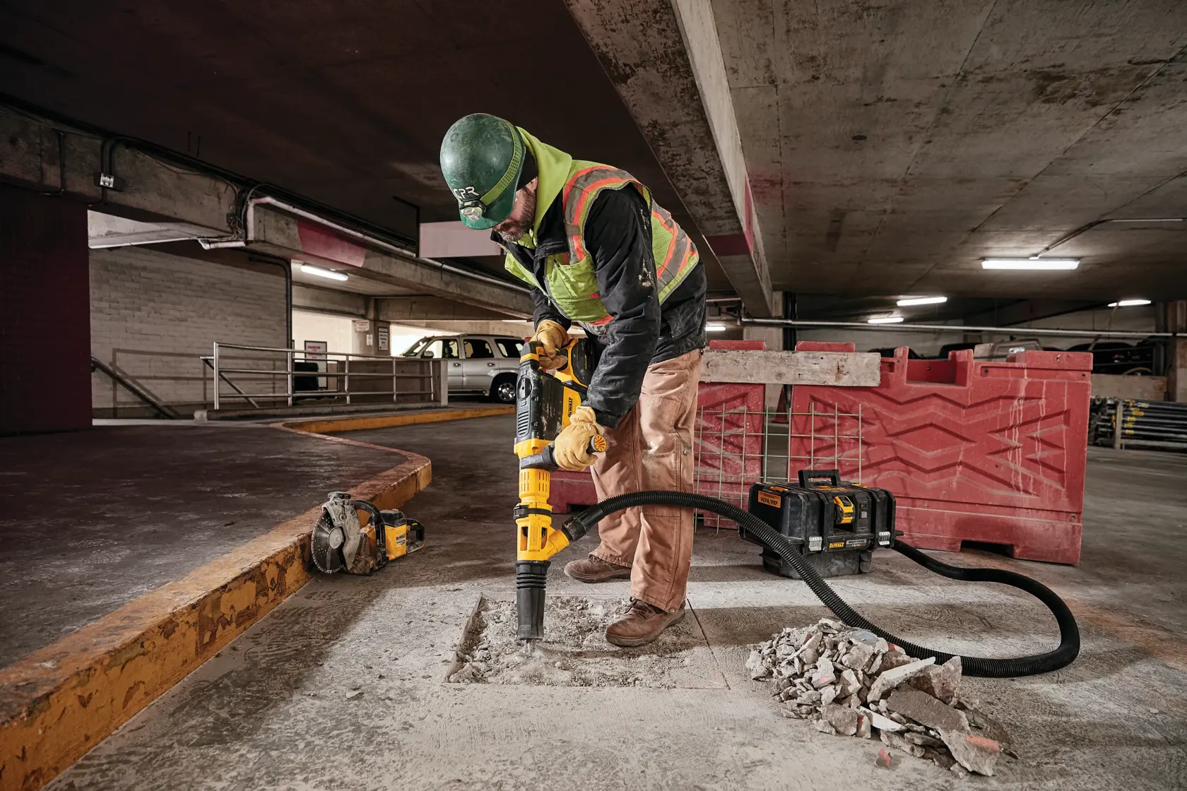 A construction worker in safety gear uses a DEWALT demolition tool with vacuum attachment to break up concrete in a parking garage. Debris is visible on the ground.