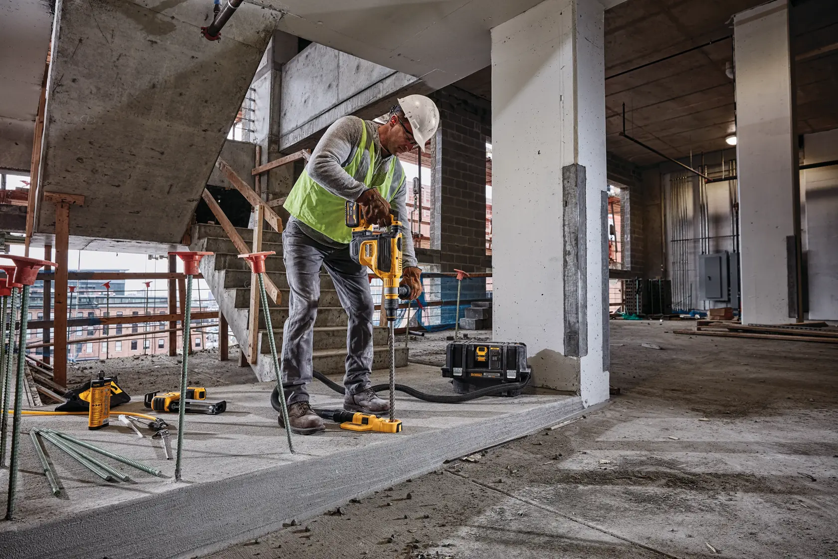 A construction worker wearing a safety vest and helmet is using a DEWALT power tool to drill into concrete indoors at a building site. DEWALT equipment and accessories are visible around the worker.