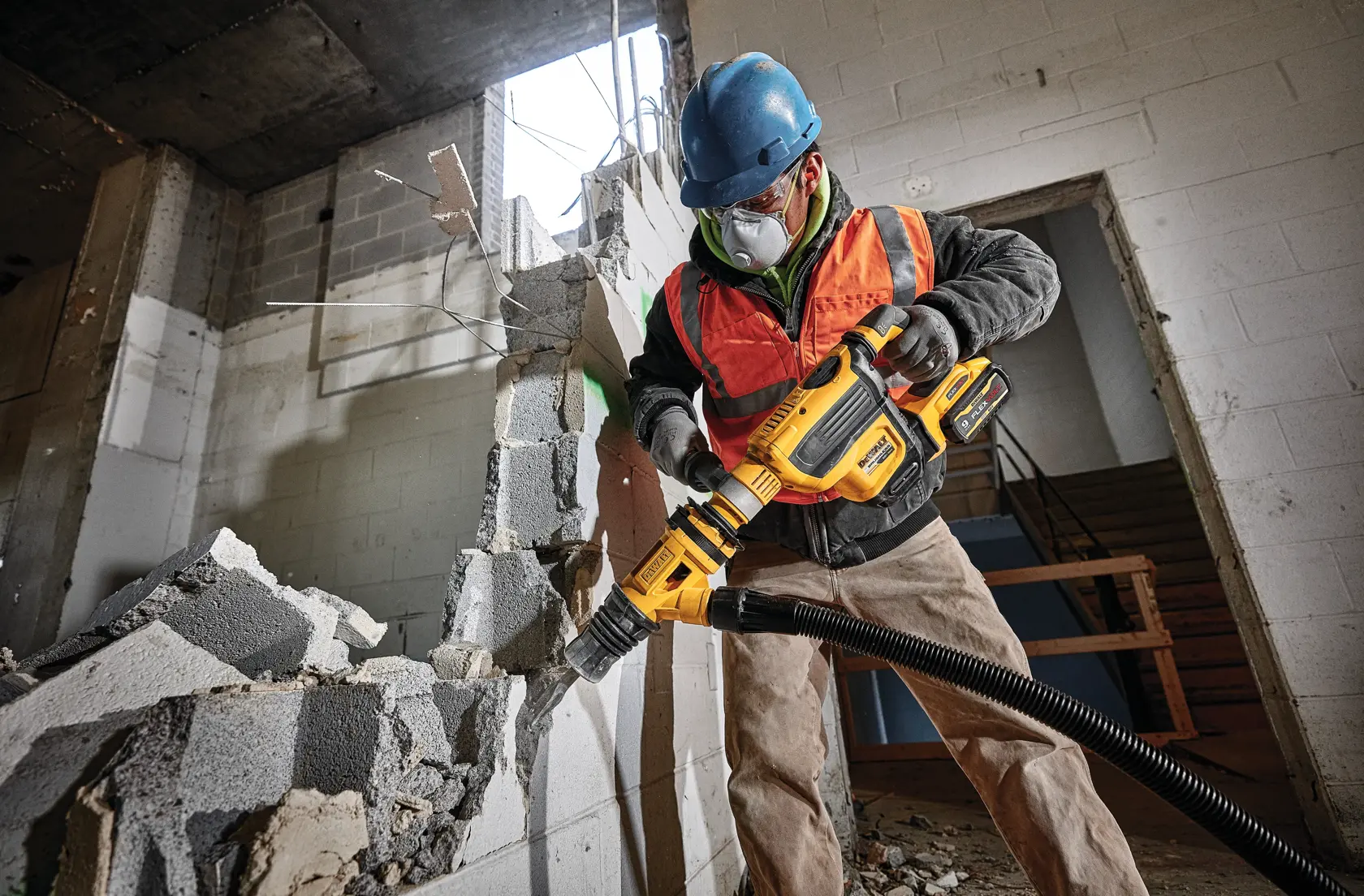 A construction worker wearing a blue hard hat and orange safety vest uses a DEWALT DCV585T2 power tool with a vacuum hose to break apart a cinder block wall in an unfinished building.