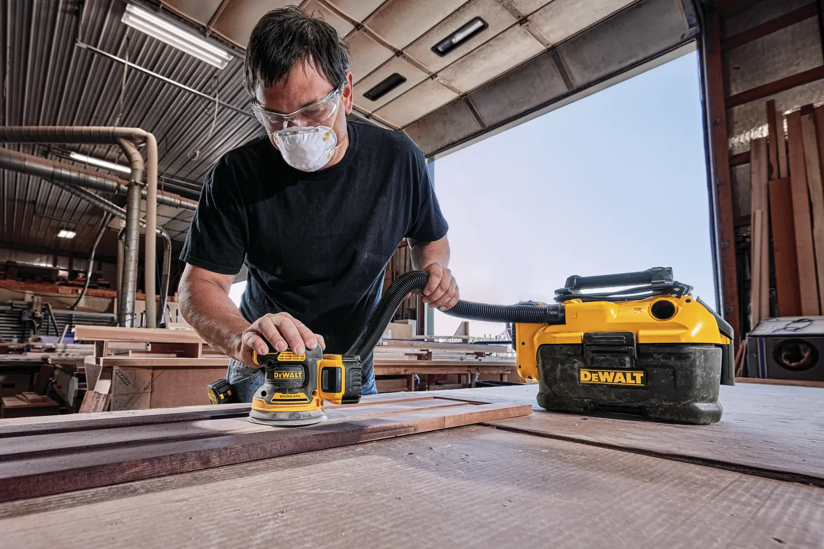 A person using a DEWALT DCW210B cordless sander in a woodworking shop, with a DEWALT vacuum attached for dust extraction. The person's face is blurred for privacy.
